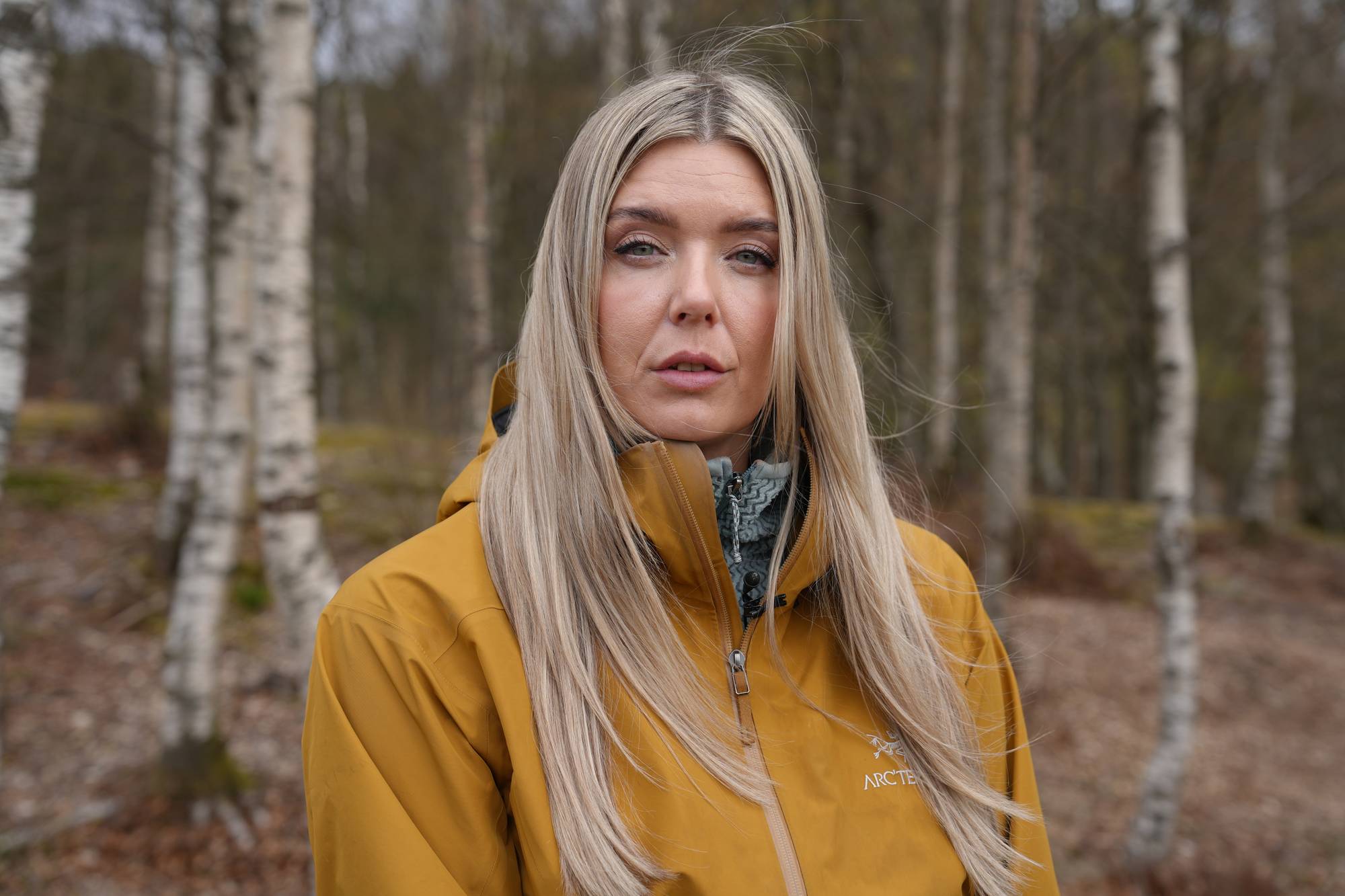 En kvinne med blondt hår og gul jakke står foran en skogkledd bakgrunn. Hun ser inn i kameraet. (Bildebeskrivelsen er laget av en KI-tjeneste)