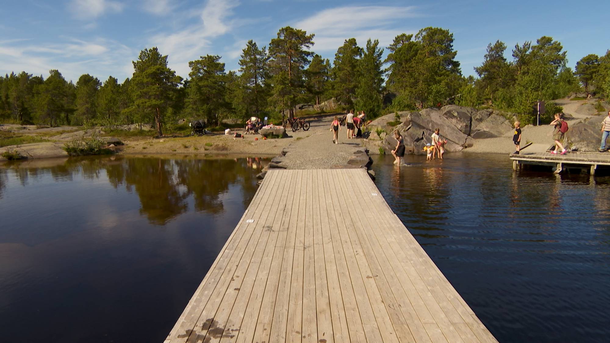 Folk oppholder seg ved en badeplass med trebrygge, svaberg og skog i bakgrunnen. (Bildebeskrivelsen er laget av en KI-tjeneste)