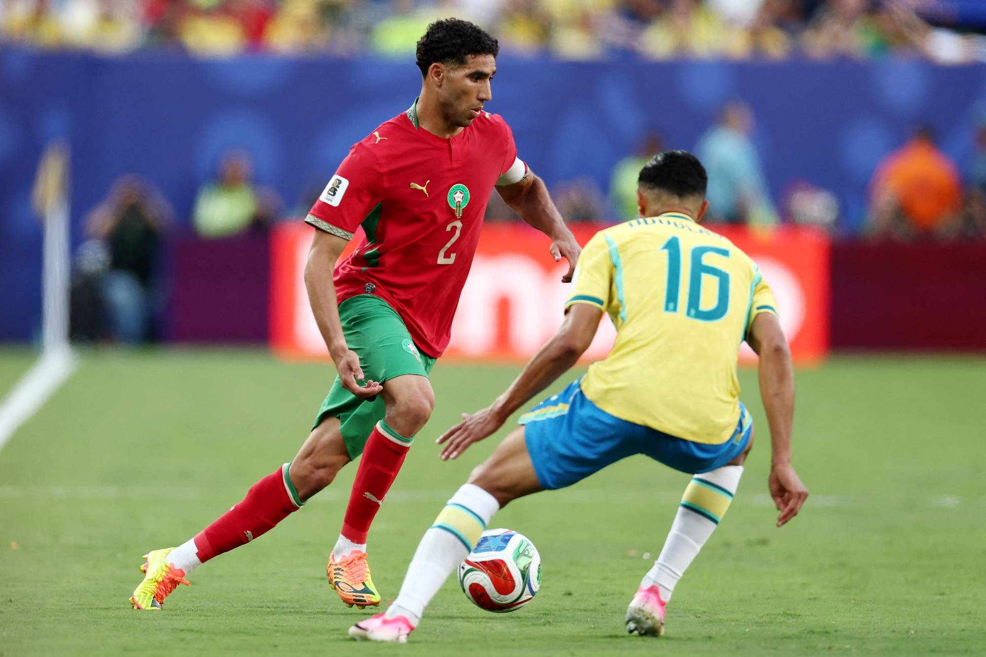 Fotballspiller fra Marokko i duell med en motspiller fra Brasil på en gressbane. 