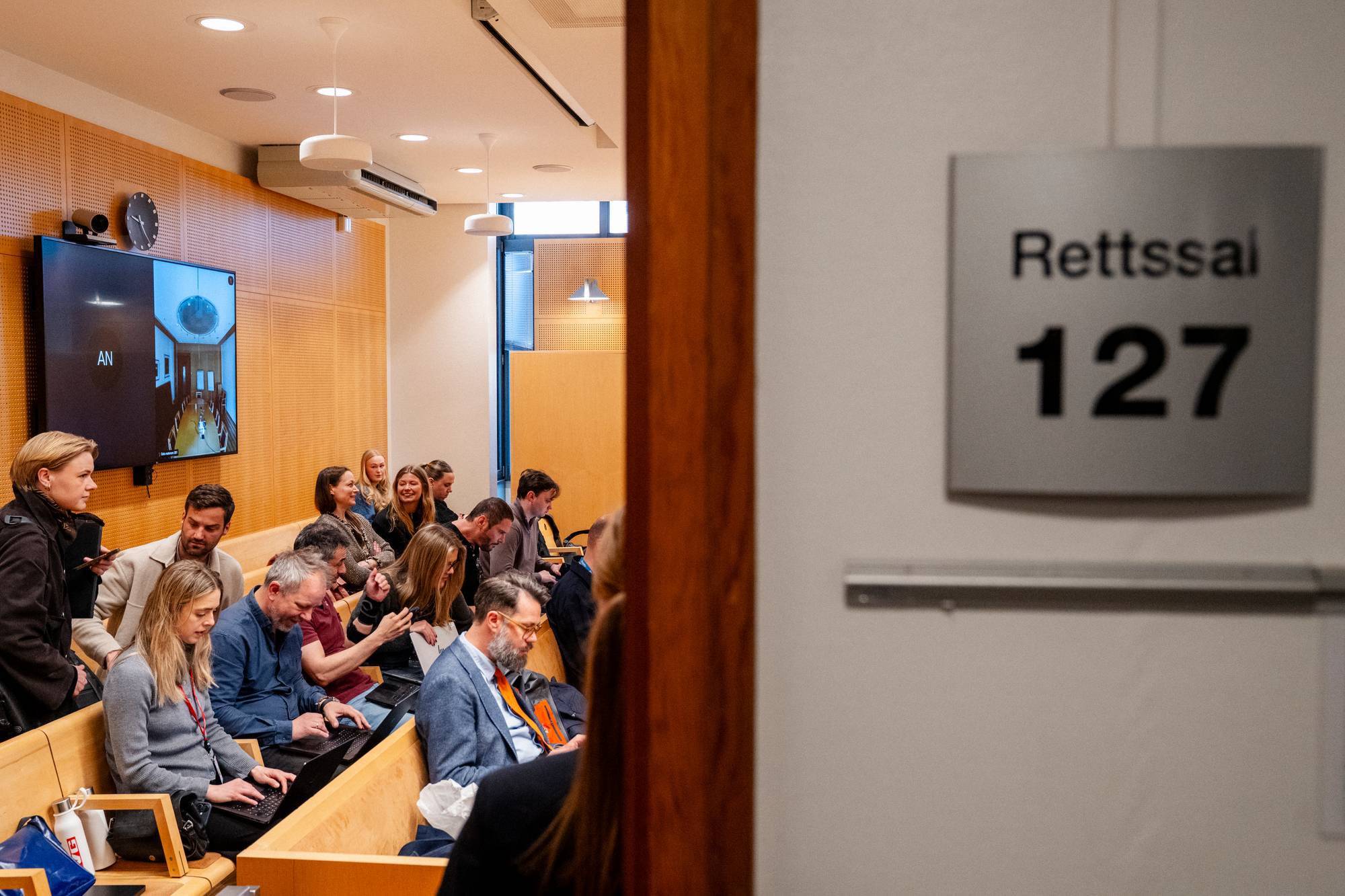 Folk sitter på benker i en rettssal. En kvinne i forgrunnen ser til venstre. En skjerm viser et bilde av en korridor. Et skilt på veggen viser teksten "Rettsal 127". (Bildebeskrivelsen er laget av en KI-tjeneste)