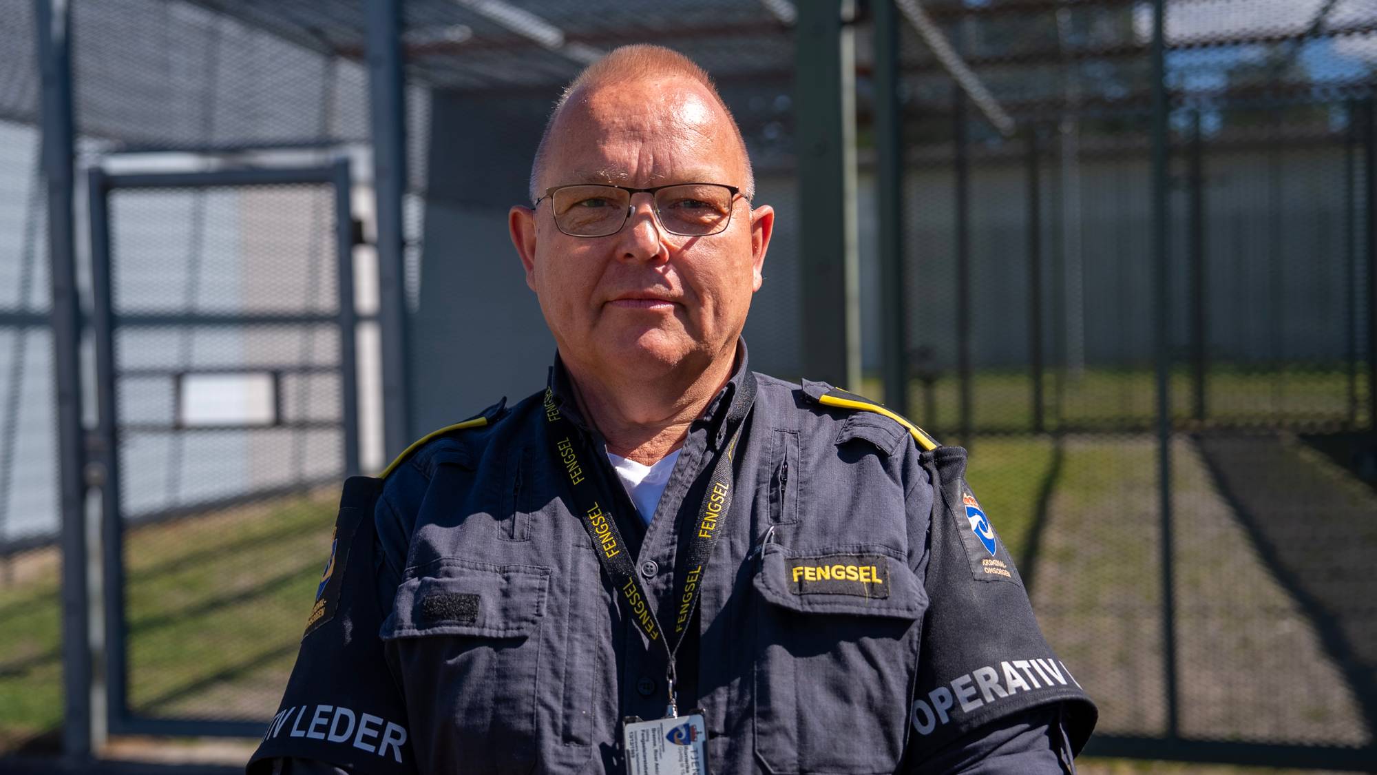Fengselsbetjent i uniform står foran et inngjerdet område utendørs. 