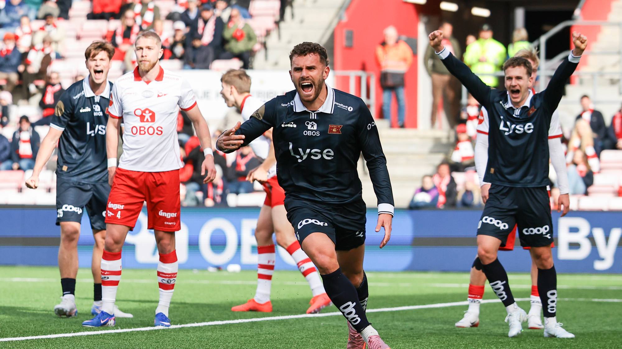 En fotballspiller jubler mens lagkamerater ser på. Spilleren er kledd i mørkeblå drakt med lyseblå detaljer. Bakgrunnen er tribunene på en fotballstadion med publikum. Flere spillere i røde og hvite drakter er også synlige på banen. (Bildebeskrivelsen er laget av en KI-tjeneste)