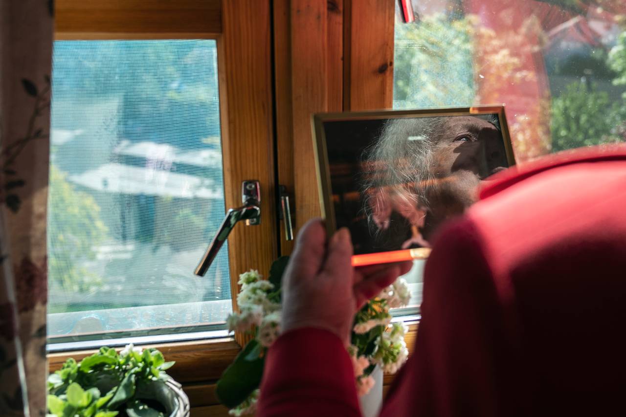Kvinne i rød jakke ser på et innrammet bilde i vinduet. I refleksjonen på bildet ser man tårer i øynene hennes.