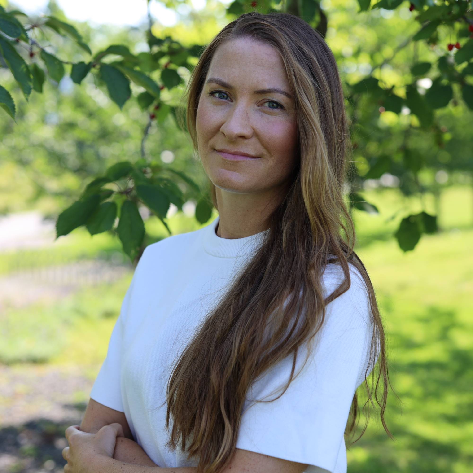 En kvinne med langt brunt hår som smiler lett mot kamera. Hun har på seg en hvit tskjorte og står utendørs hvor det er grønt gress og trær rundt henne.