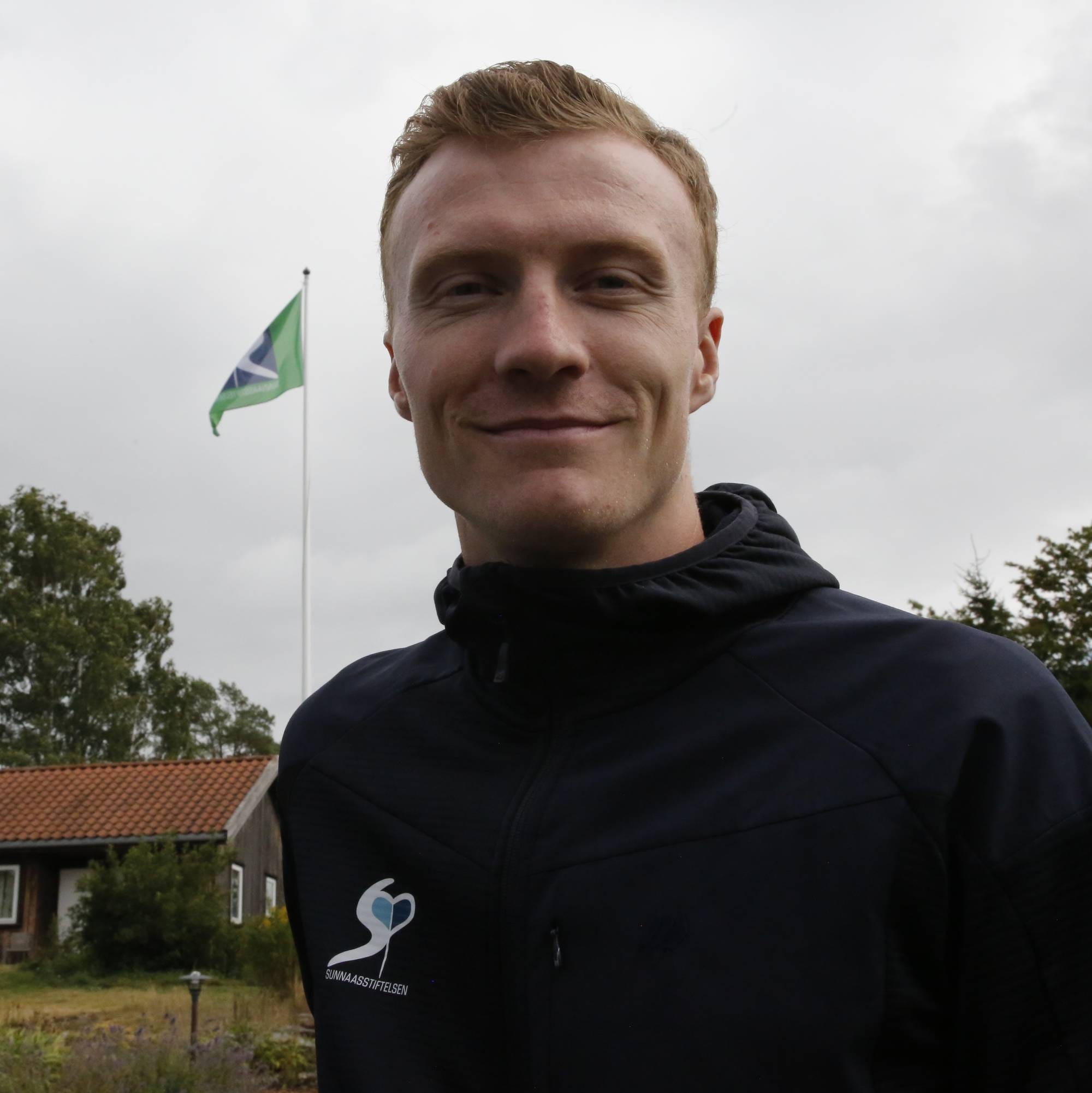 Mann med rødt hår og mørk jakke utendørs, med en bygning og flagg i bakgrunnen. (Bildebeskrivelsen er laget av en KI-tjeneste)