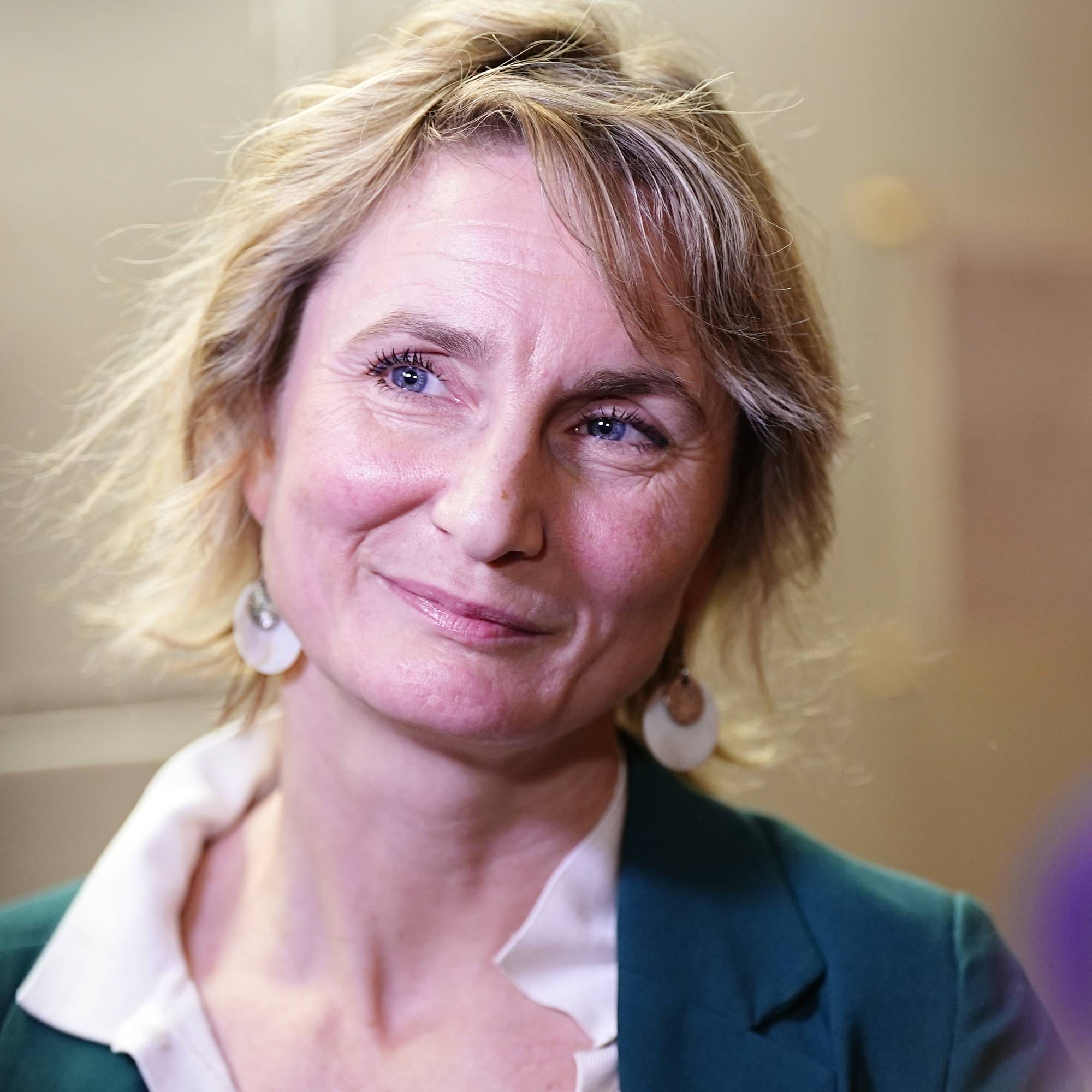 Marthe Hammer med kort, lys hår står i fokus. Hun har på seg en grønn jakke med hvit krage. Bakgrunnen er uskarp, men viser indre lokaler. Atmosfæren tyder på en formell setting.