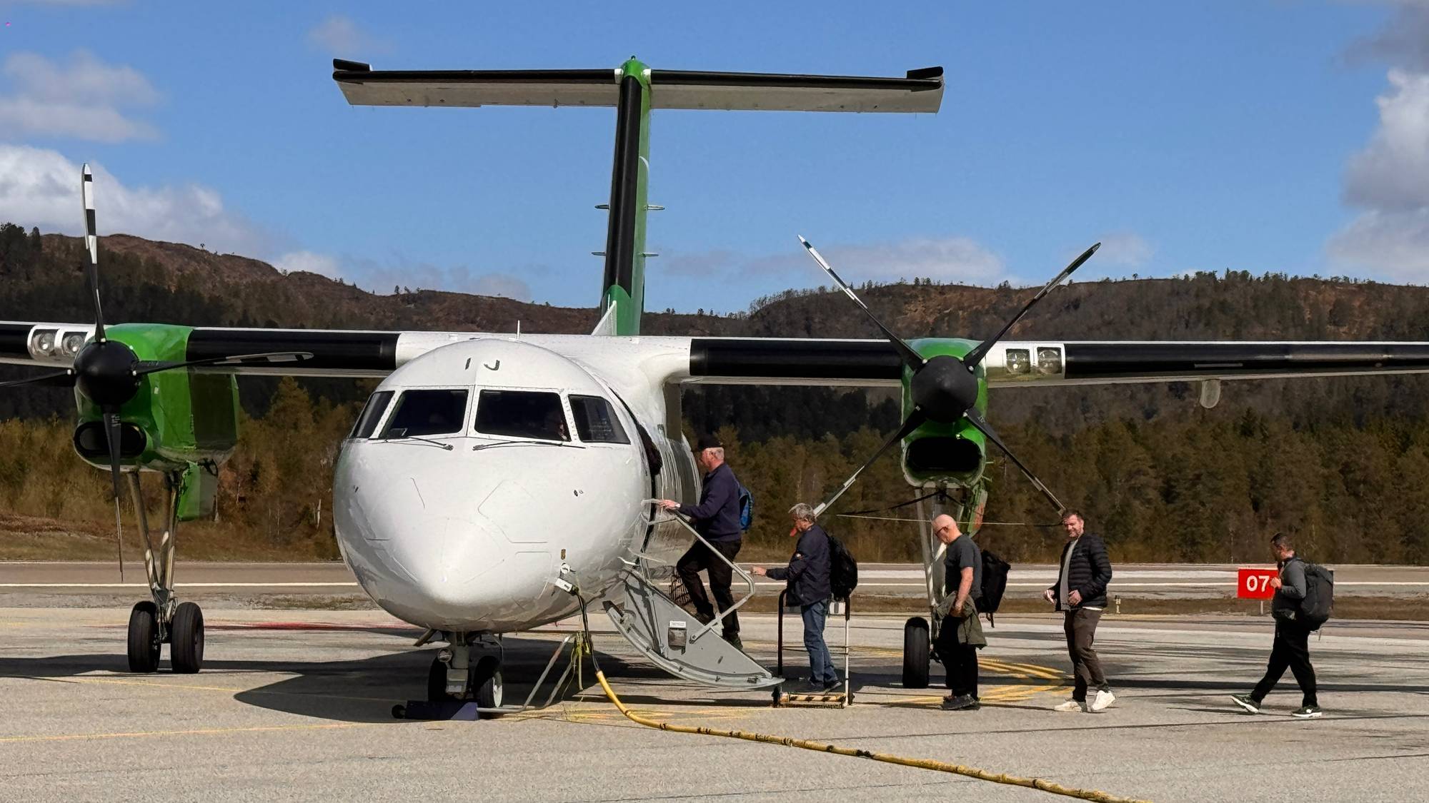 Passasjerer går om bord i et småfly på en rullebane. Flyet er hvitt med grønne motordeksel og hale. Bak flyet ligger et skogkledd landskap. Himmelen er blå med hvite skyer. (Bildebeskrivelsen er laget av en KI-tjeneste)