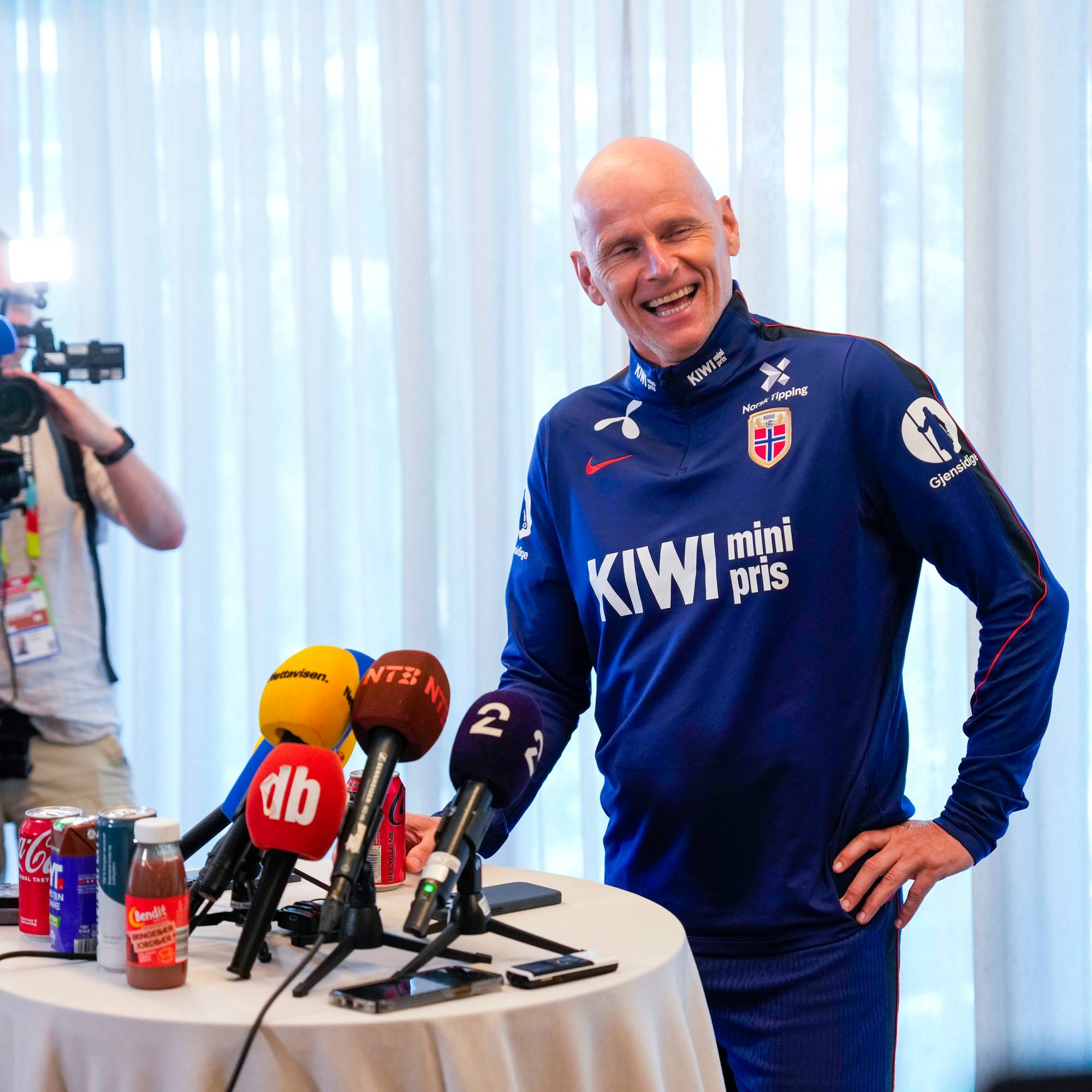 Landslagstrener Ståle Solbakken ler ved et bord med mikrofoner på en pressekonferanse. (Bildebeskrivelsen er laget av en KI-tjeneste)