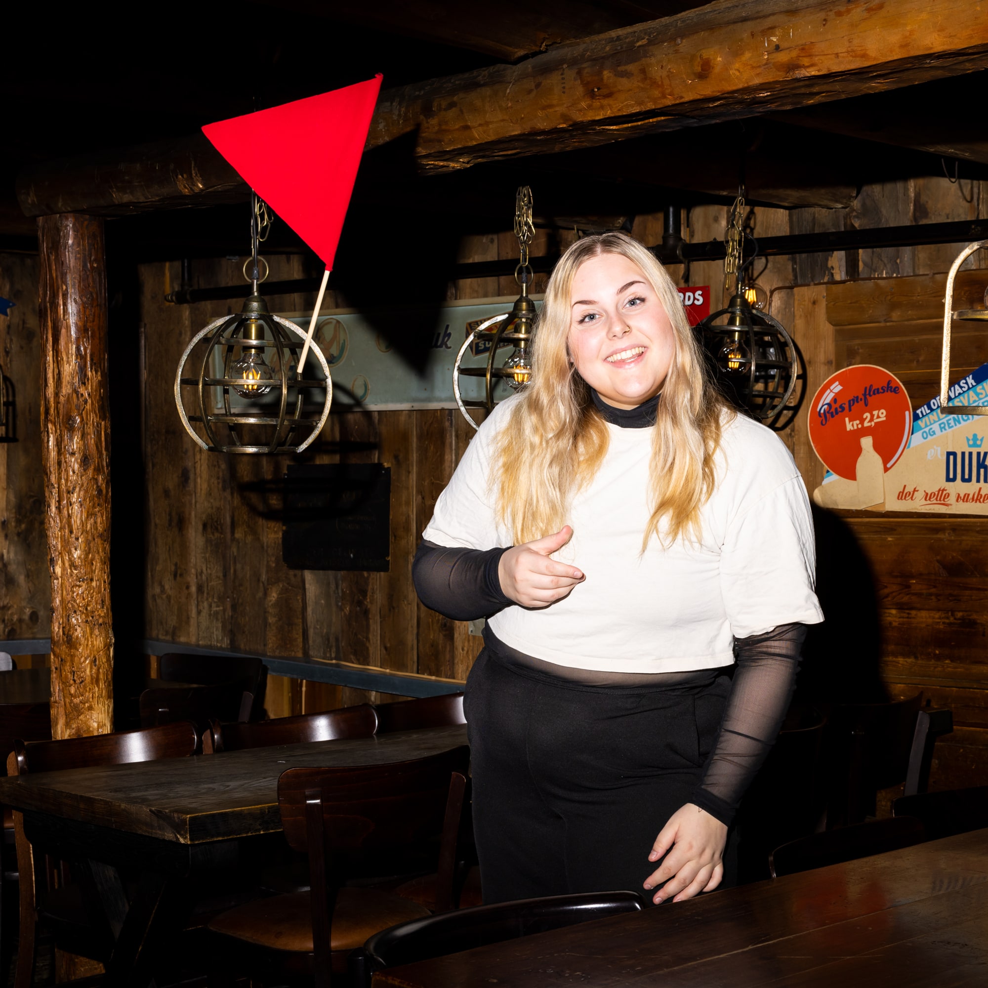 Cathrine er på en pub i Trondheim. Interiøret er av tre. Hun smiler stort mens hun kaster et rødt flagg i luften. 