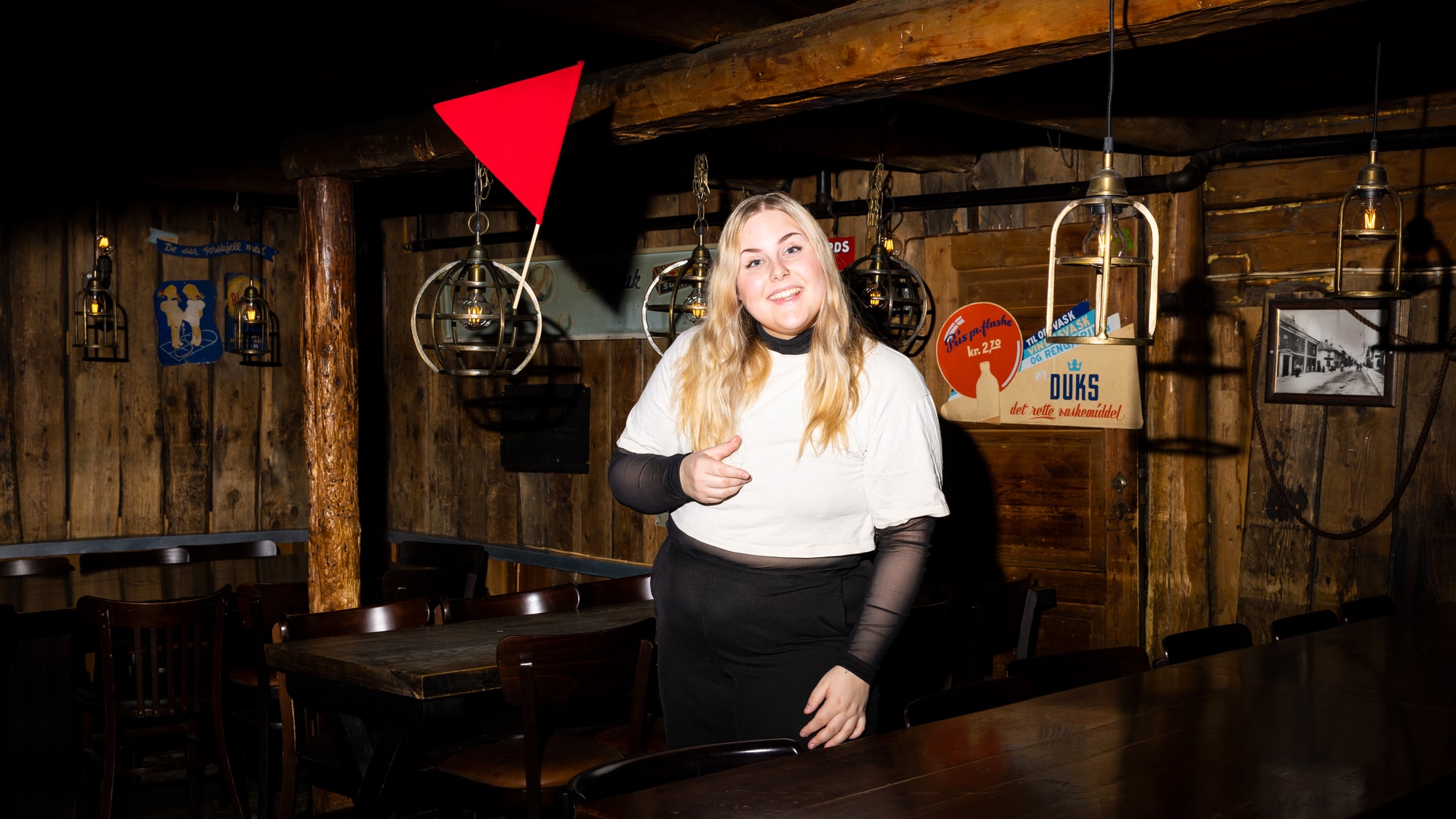 Cathrine er på en pub i Trondheim. Interiøret er av tre. Hun smiler stort mens hun kaster et rødt flagg i luften. 