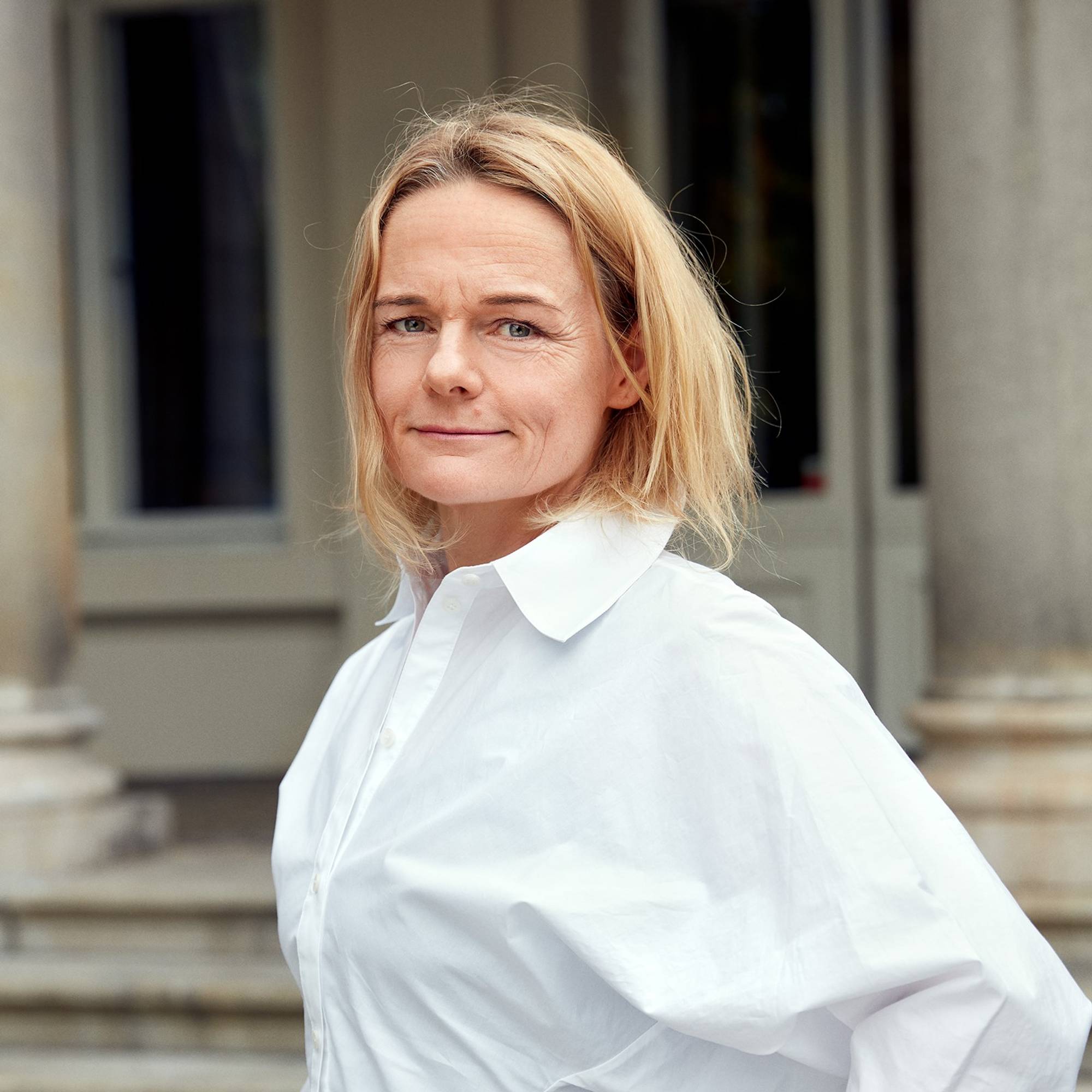 En person står på en trapp foran en bygning. Hun har på seg en hvit skjorte med store ermer og har blondt hår. Omgivelsene viser en bygning med søyler og vinduer. Det ser ut til å være en rolig dag med naturlig lys. (Bildebeskrivelsen er laget av en KI-tjeneste)