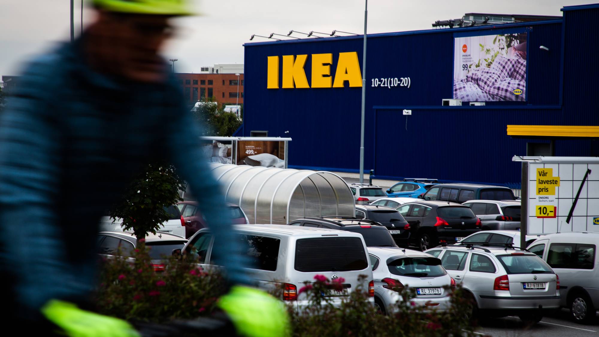 En uskarpt syklist med grønn hjelm passerer en stor IKEA-butikk. Bildet viser en full parkeringsplass foran den blå bygningen. På fasaden er det et stort gult IKEA-skilt og et bilde med tekst. En stor lys reklameplakat viser tilbudspris. (Bildebeskrivelsen er laget av en KI-tjeneste)