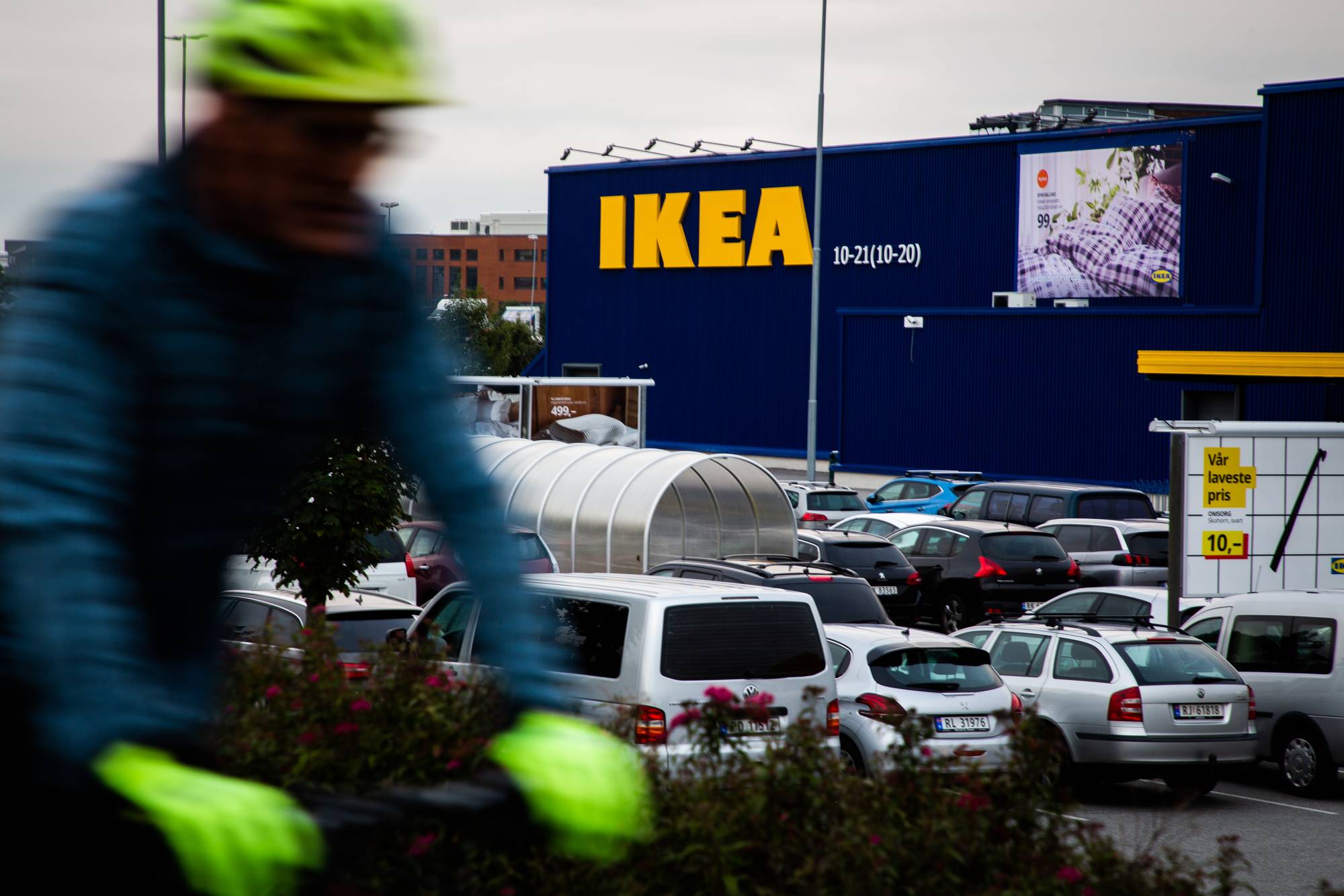 En uskarpt syklist med grønn hjelm passerer en stor IKEA-butikk. Bildet viser en full parkeringsplass foran den blå bygningen. På fasaden er det et stort gult IKEA-skilt og et bilde med tekst. En stor lys reklameplakat viser tilbudspris. (Bildebeskrivelsen er laget av en KI-tjeneste)