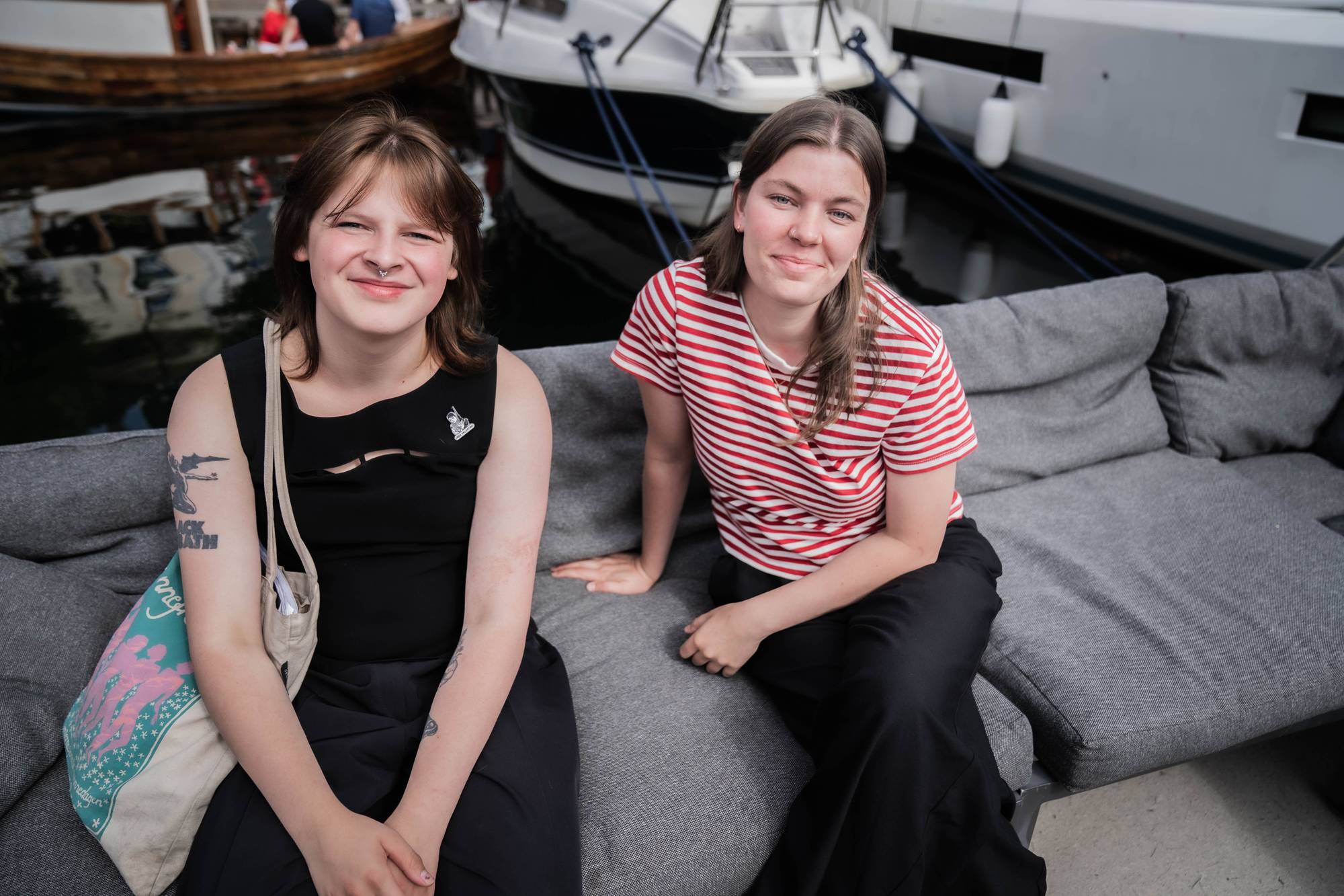 To unge kvinner sitter på en grå sofa ved en småbåthavn. 