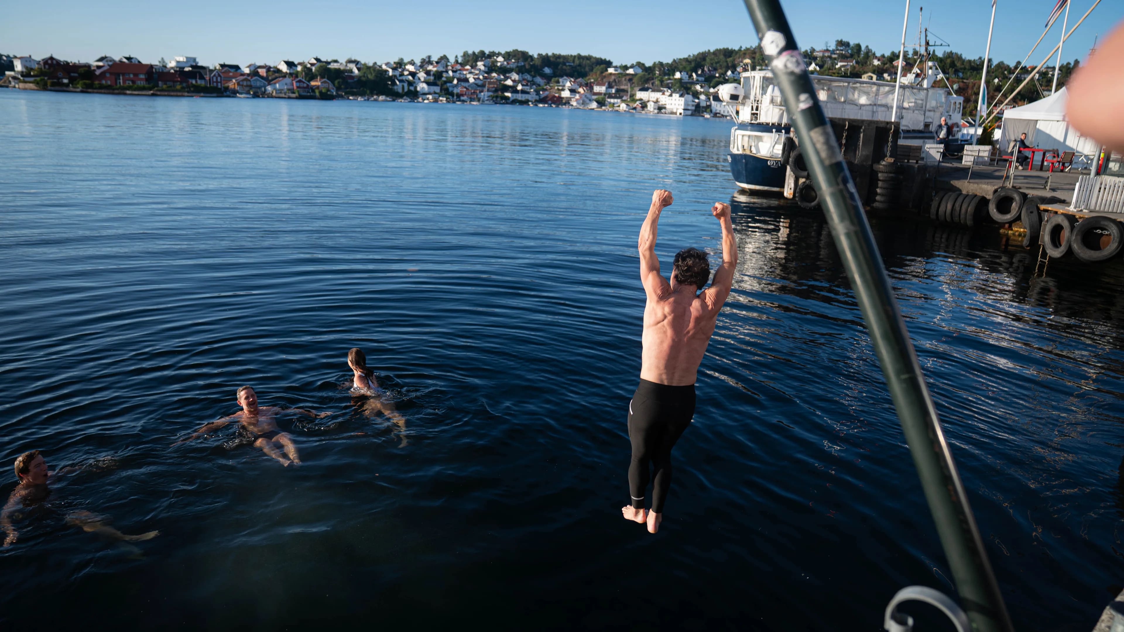 Person hopper i sjøen fra kai mens tre personer bader i nærheten i en havn.