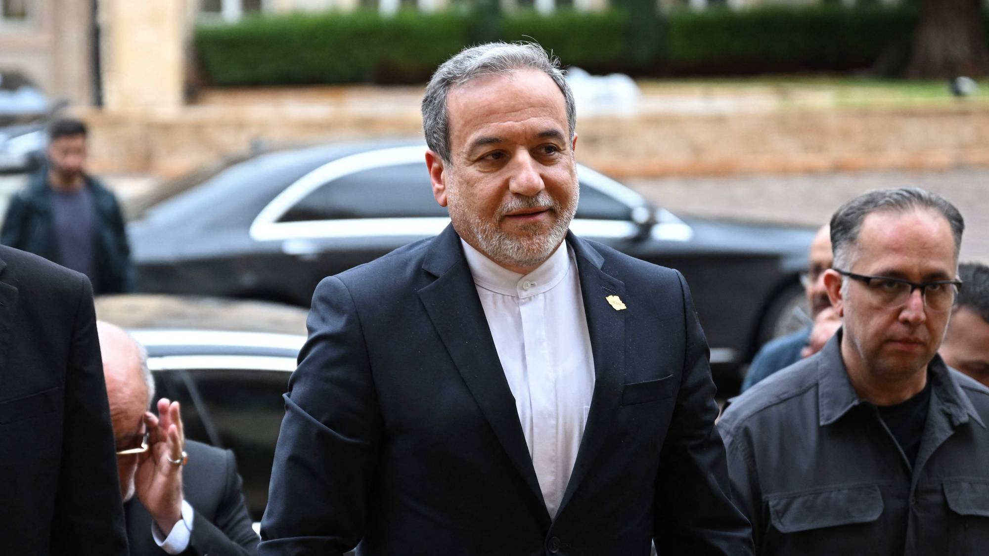 Flere menn i dress går ut av en bygning, mens en svart bil står parkert bak dem. En av mennene bærer et insign som indikerer at han er del av den iranske delegasjonen. Bygningen har en klassisk fasade med buede vinduer og er omgitt av trær. (Bildebeskrivelsen er laget av en KI-tjeneste)
