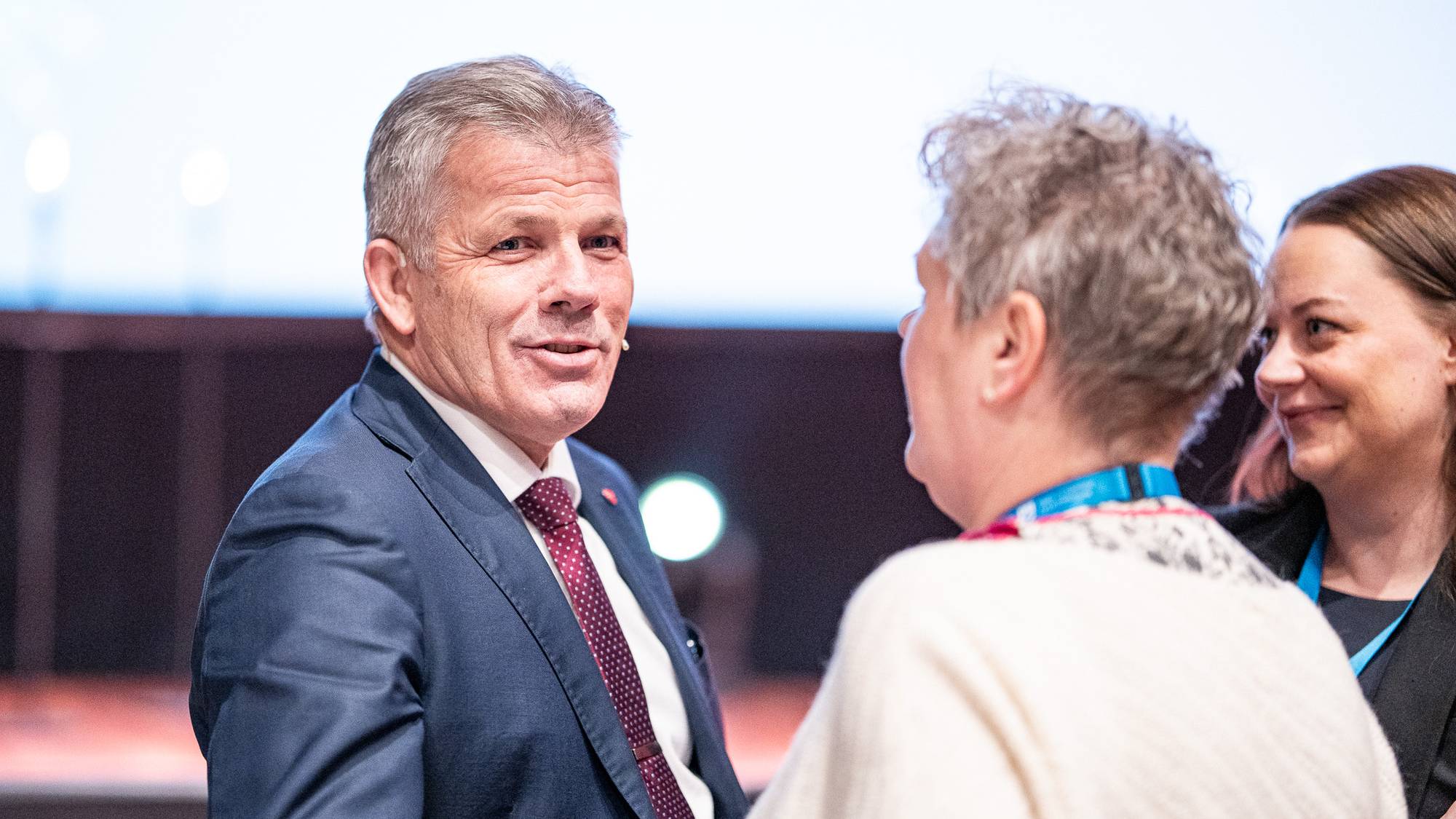 En mann i dress står og snakker med en kvinne, som har på seg en lys genser. En annen kvinne, delvis synlig, lytter til samtalen. Bakgrunnen er en konferansesal med dempet belysning og en stor skjerm med lys. Bildeutsnittet fokuserer på de tre personene i interaksjon. (Bildebeskrivelsen er laget av en KI-tjeneste)