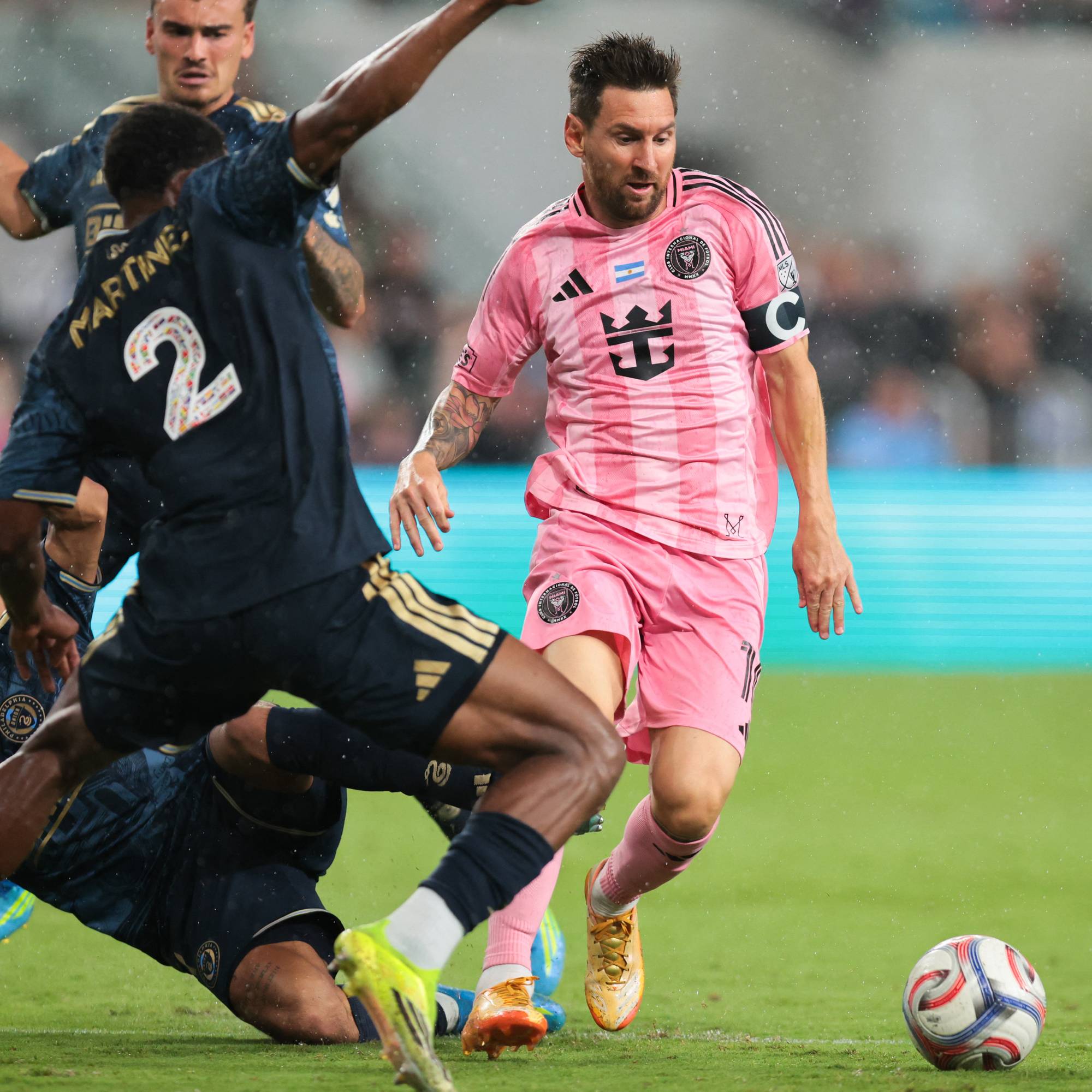 Lionel Messi i rosa drakt løper med ballen. Fotballspillere i mørkeblå drakt tackler i forgrunnen. En dommer i grønn trøye løper bak i bildet. (Bildebeskrivelsen er laget av en KI-tjeneste)
