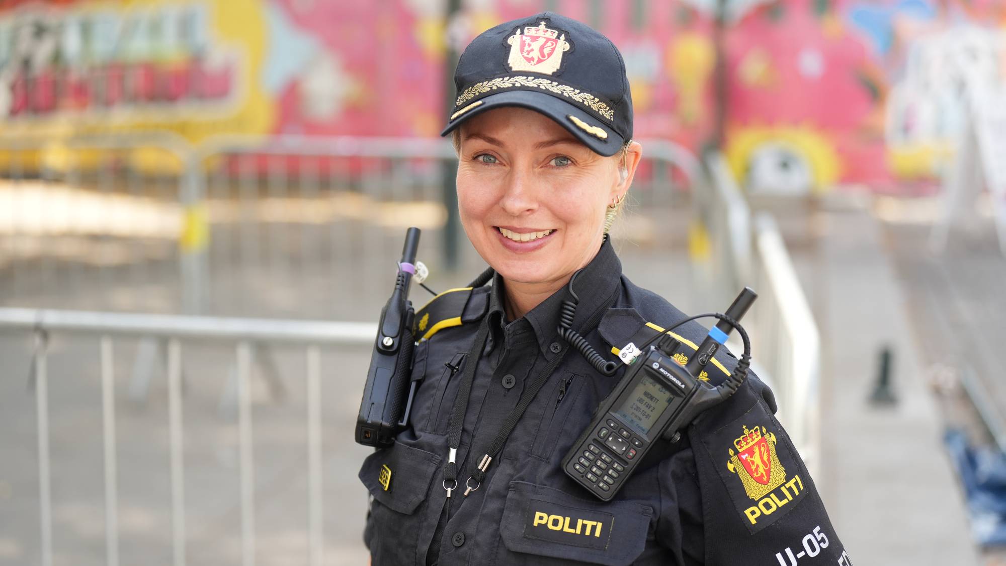 En kvinnelig politibetjent i uniform smiler foran et metallgjerde utendørs. (Bildebeskrivelsen er laget av en KI-tjeneste)