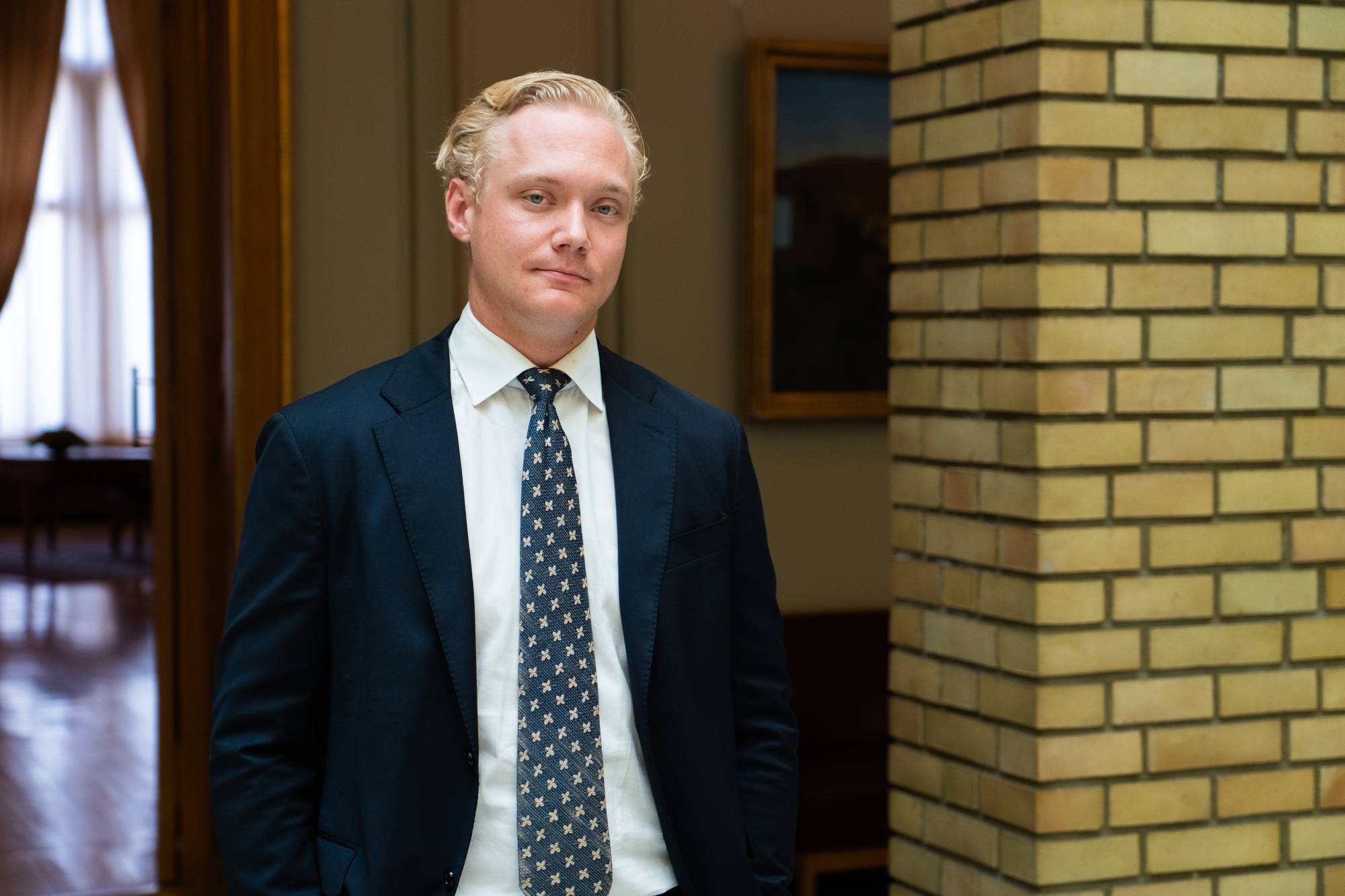 En mann med lyst hår og dress står vendt mot kameraet. Han har på seg en mørkeblå dress, hvit skjorte og et mønstret slips. Bak ham er en vegg av gule murstein. I bakgrunnen ser man et maleri på veggen og en åpen døråpning. (Bildebeskrivelsen er laget av en KI-tjeneste)