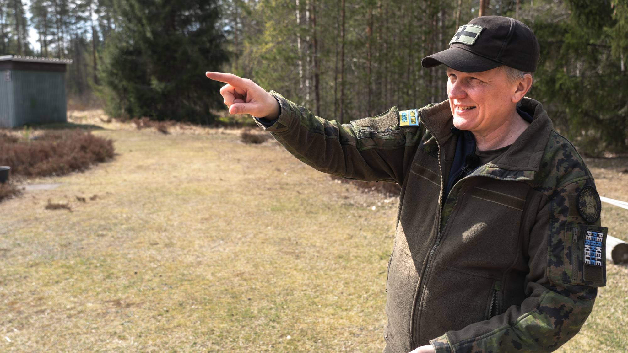 En mann i militæruniform peker mot venstre. Han har på seg en mørk caps og en mørkegrønn jakke med kamuasjemønster. I bakgrunnen er det trær og en liten bygning. (Bildebeskrivelsen er laget av en KI-tjeneste)
