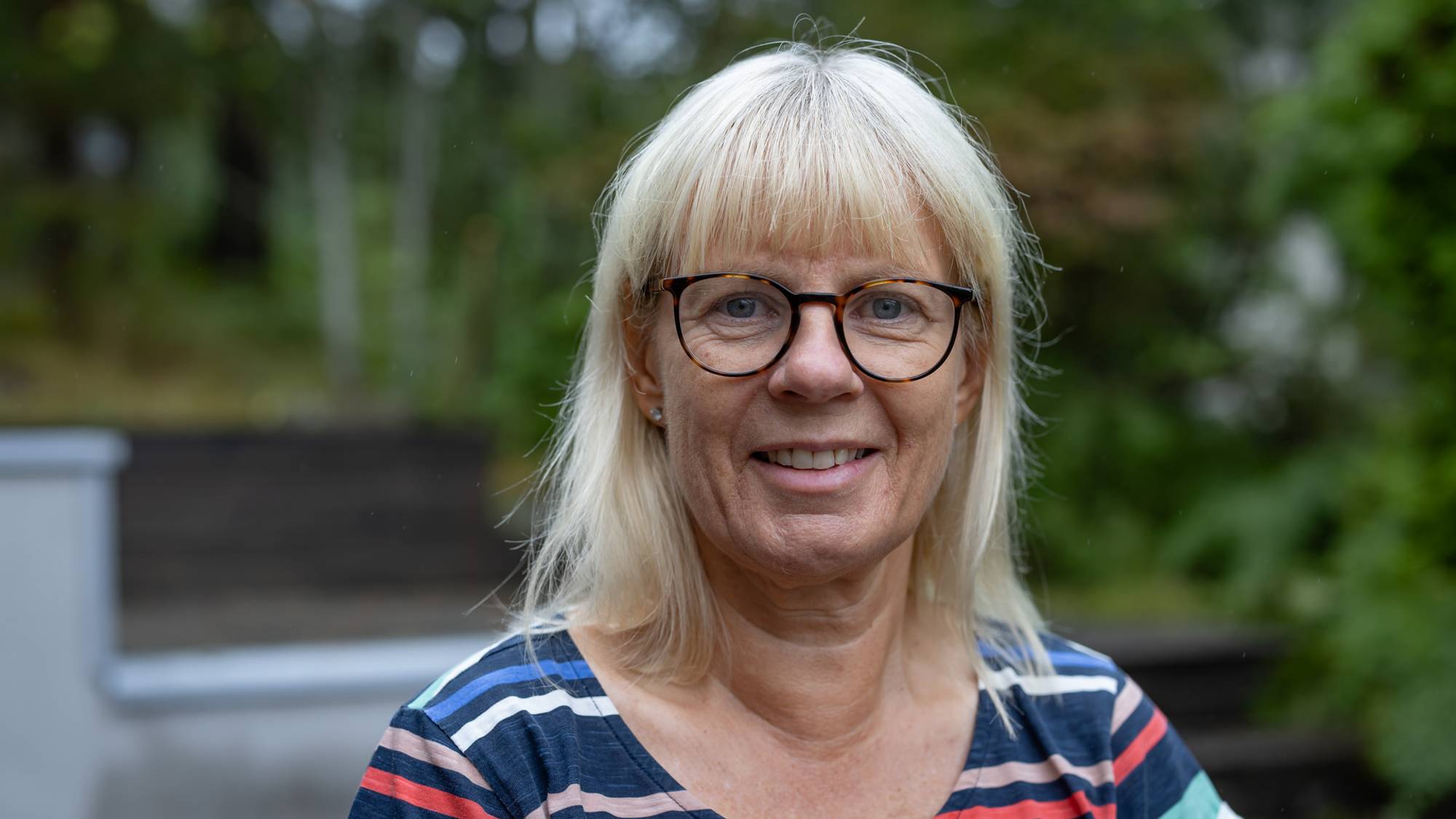 En kvinne med briller kikker mot kamera og smiler. Hun har langt lyst hår og en stripete genser på seg. I bakgrunnen ser vi trær og deler av en hage