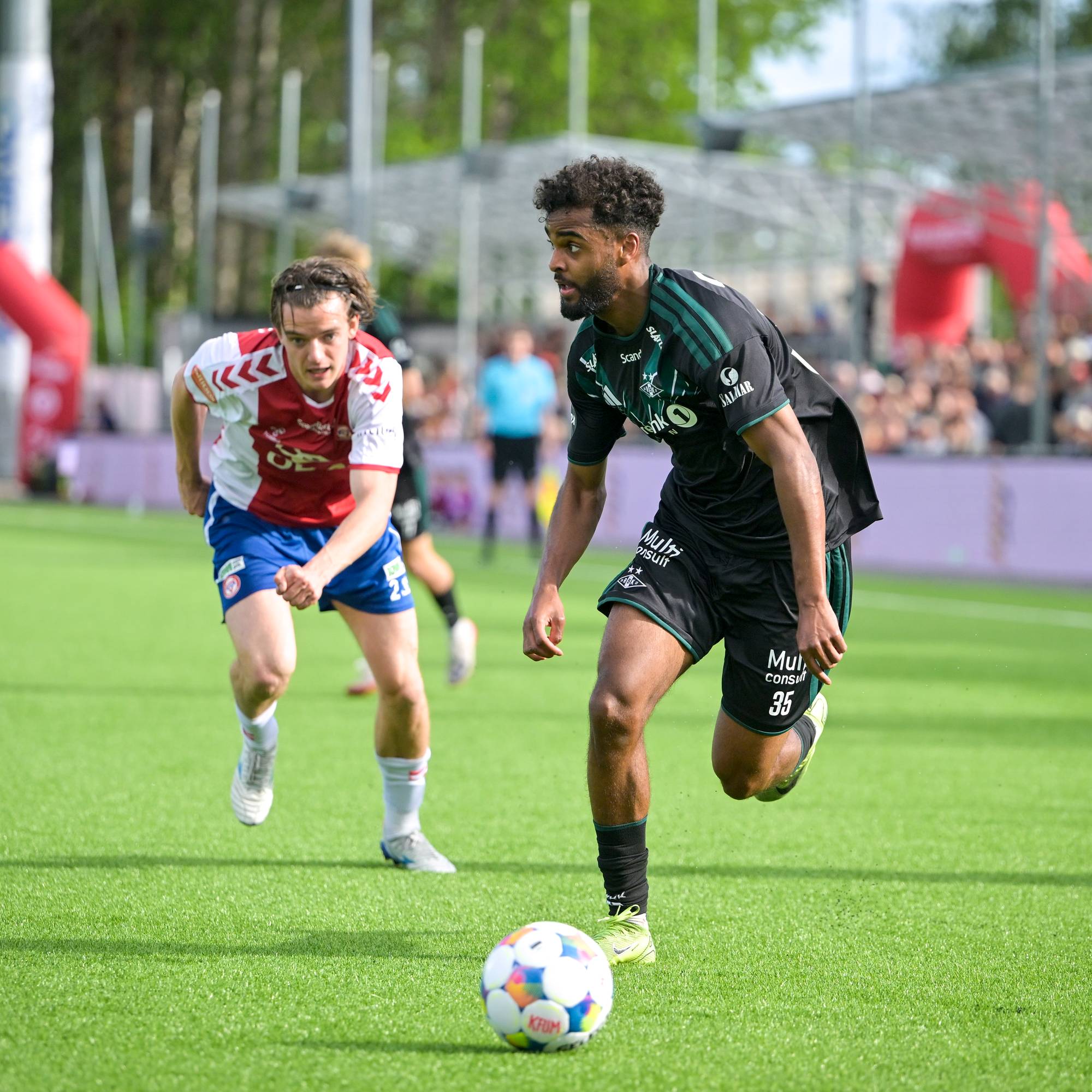 Fotballspillere i aksjon på en grønn gressmatte. En spiller i svart drakt med nummer 35 presser ballen fremover. En annen spiller i rød og hvit drakt løper ved siden av. Publikum sitter på tribunen i bakgrunnen. (Bildebeskrivelsen er laget av en KI-tjeneste)