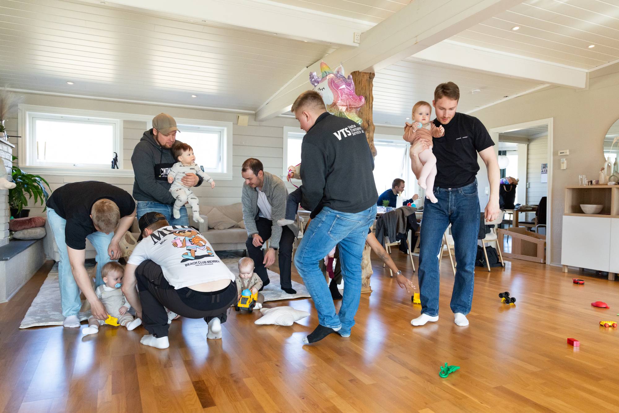 En gruppe fedre samler babyene sine til gruppebilde inne i en stor stue, med leker på gulvet rundt dem