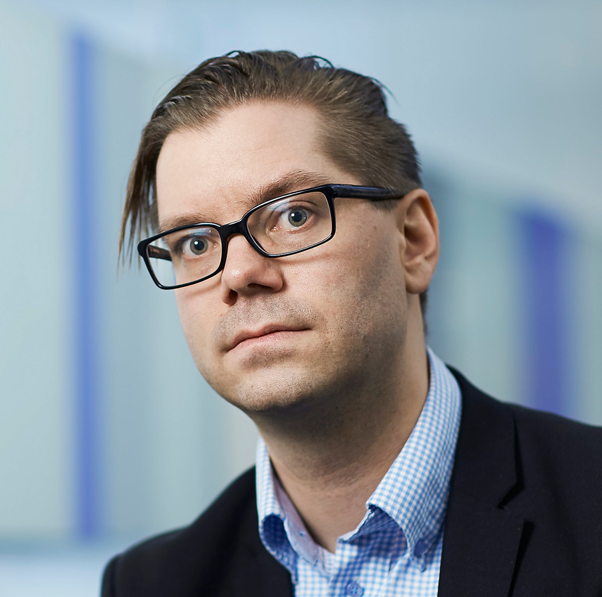 En mann med sleik, briller og noen få skjeggstubber ser inn i kameraet. Han har på seg en stripete, blå skjorte og dressjakke. 