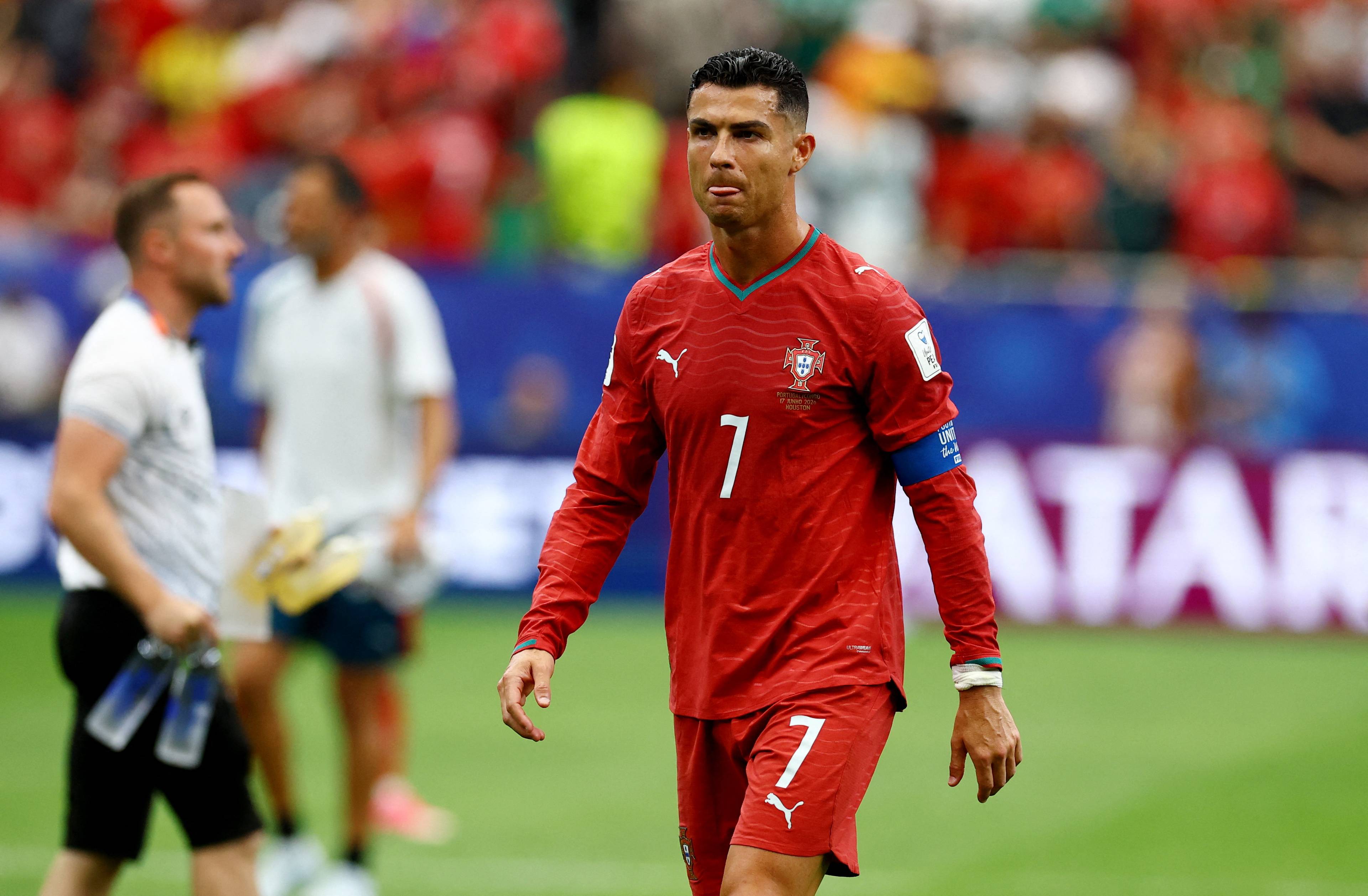 Cristiano Ronaldo i rød drakt. Han har nummer 7 på ryggen og på shortsen. Bak ham står et par andre personer. Publikum sitter på tribunen i bakgrunnen. (Bildebeskrivelsen er laget av en KI-tjeneste)