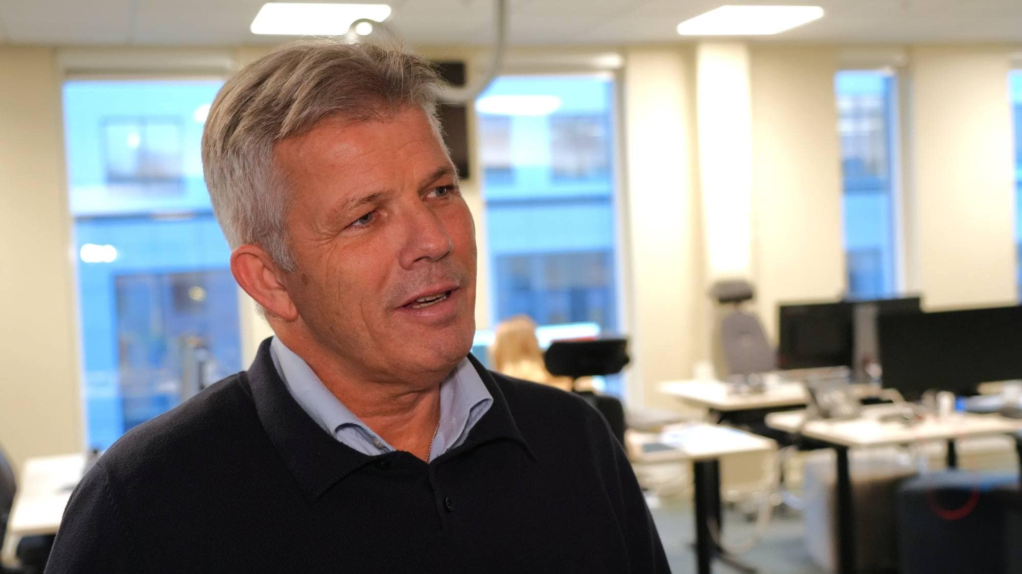 En mann i svart genser og blå skjorte står i et kontorlokale.