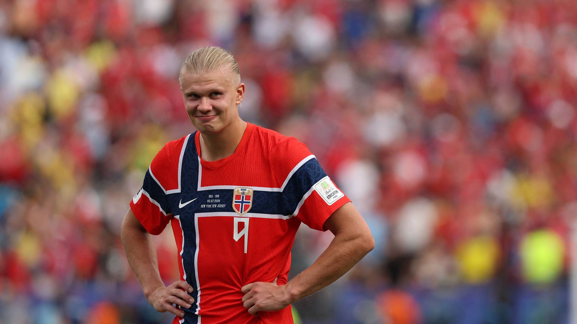 Erling Braut Haaland i norsk landslagsdrakt på en fotballbane med publikum i bakgrunnen. (Bildebeskrivelsen er laget av en KI-tjeneste)