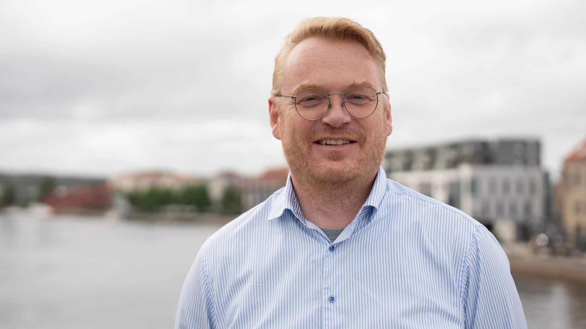 En mann med rødt hår og briller smiler mot kamera. Han har på seg en lyseblå skjorte med striper. I bakgrunnen er det en uskarp bysilhuett med bygninger og vann. (Bildebeskrivelsen er laget av en KI-tjeneste)