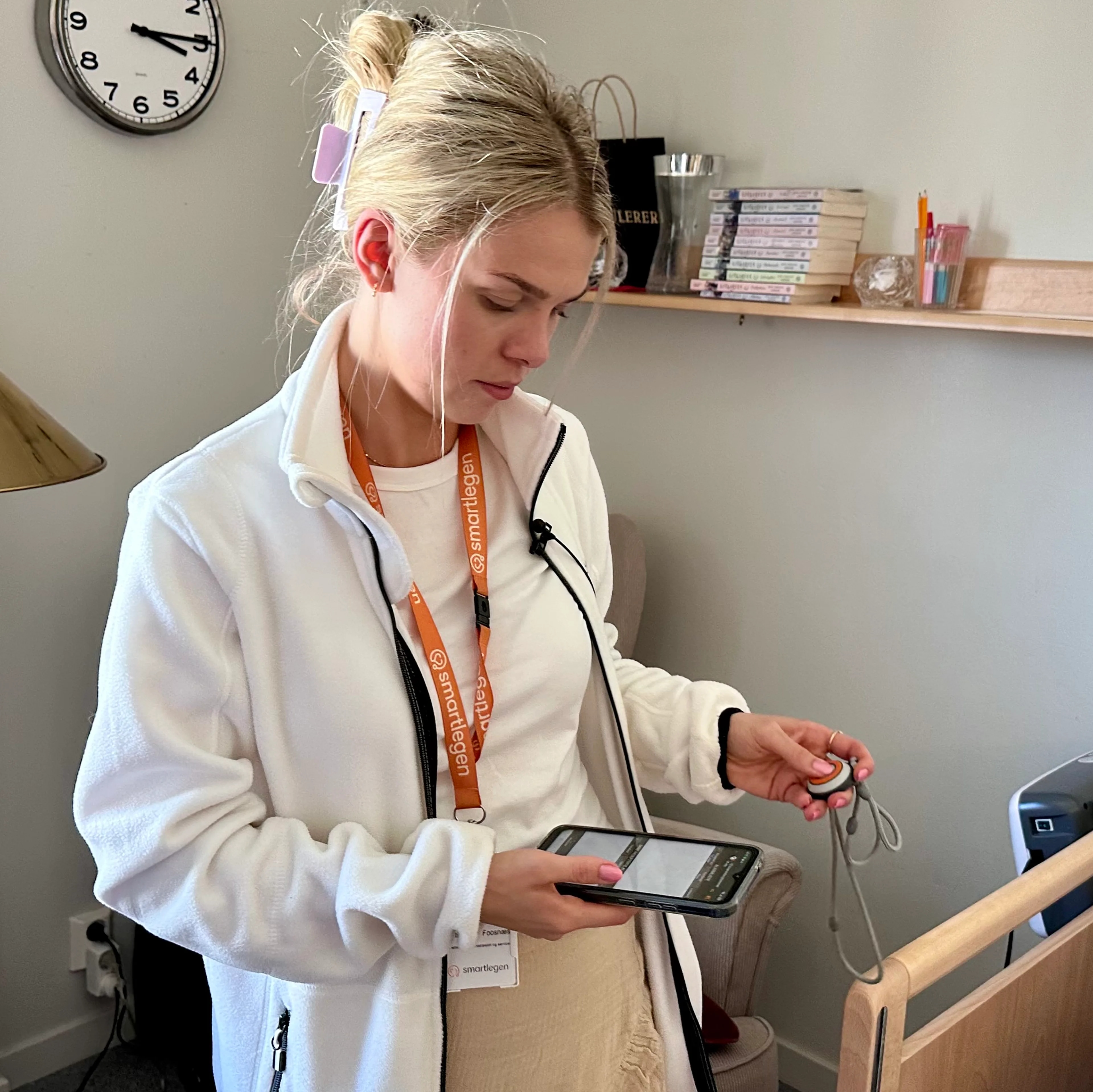 Kvinne i hvit jakke med ID-bånd holder en mobiltelefon og en trygghetsalarm i et rom med seng. (Bildebeskrivelsen er laget av en KI-tjeneste)