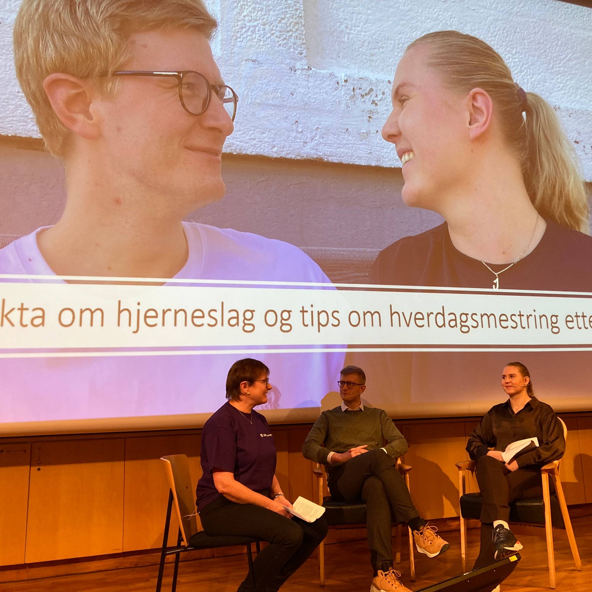 En skjerm med en mann og en dame som ser på hverandre. Under skjermen sitter et panel på tre stykker og har en samtale.