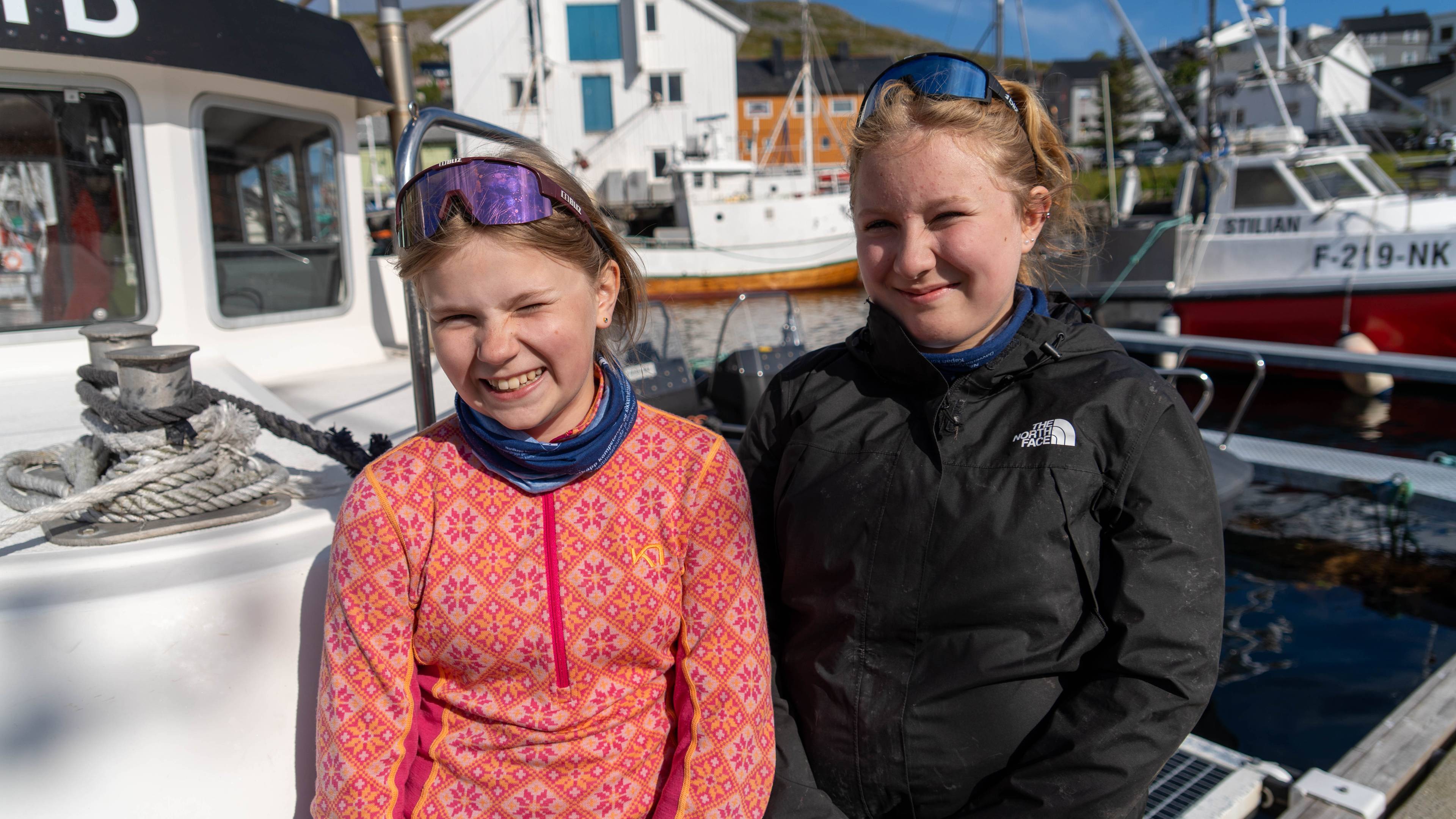 To unge jenter står foran en båt i en havn med fiskebåter i bakgrunnen. (Bildebeskrivelsen er laget av en KI-tjeneste)