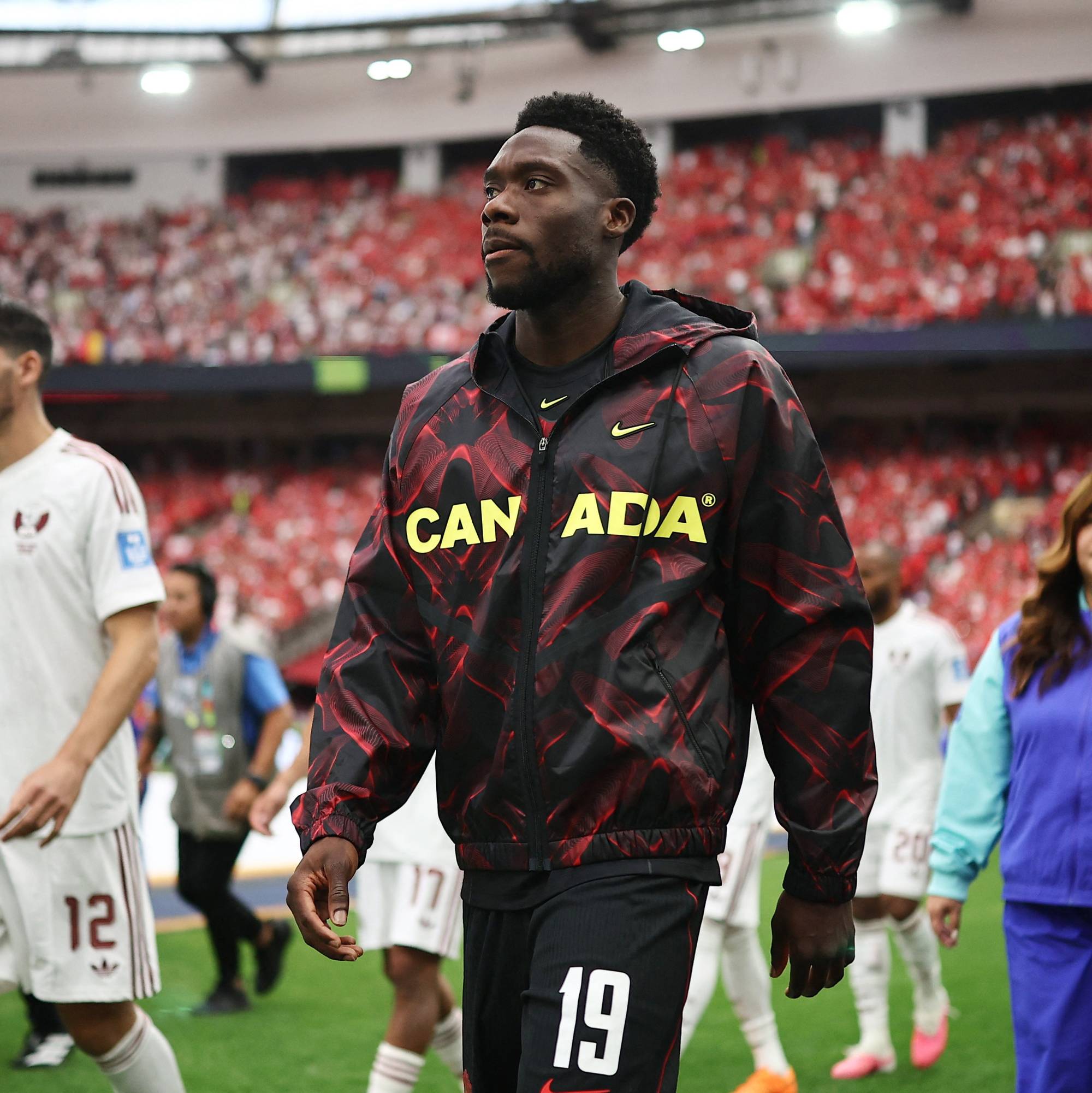 Alphonso Davies går over gressmatta på en fotballstadion ikledd Canadas treningsjakke. (Bildebeskrivelsen er laget av en KI-tjeneste)