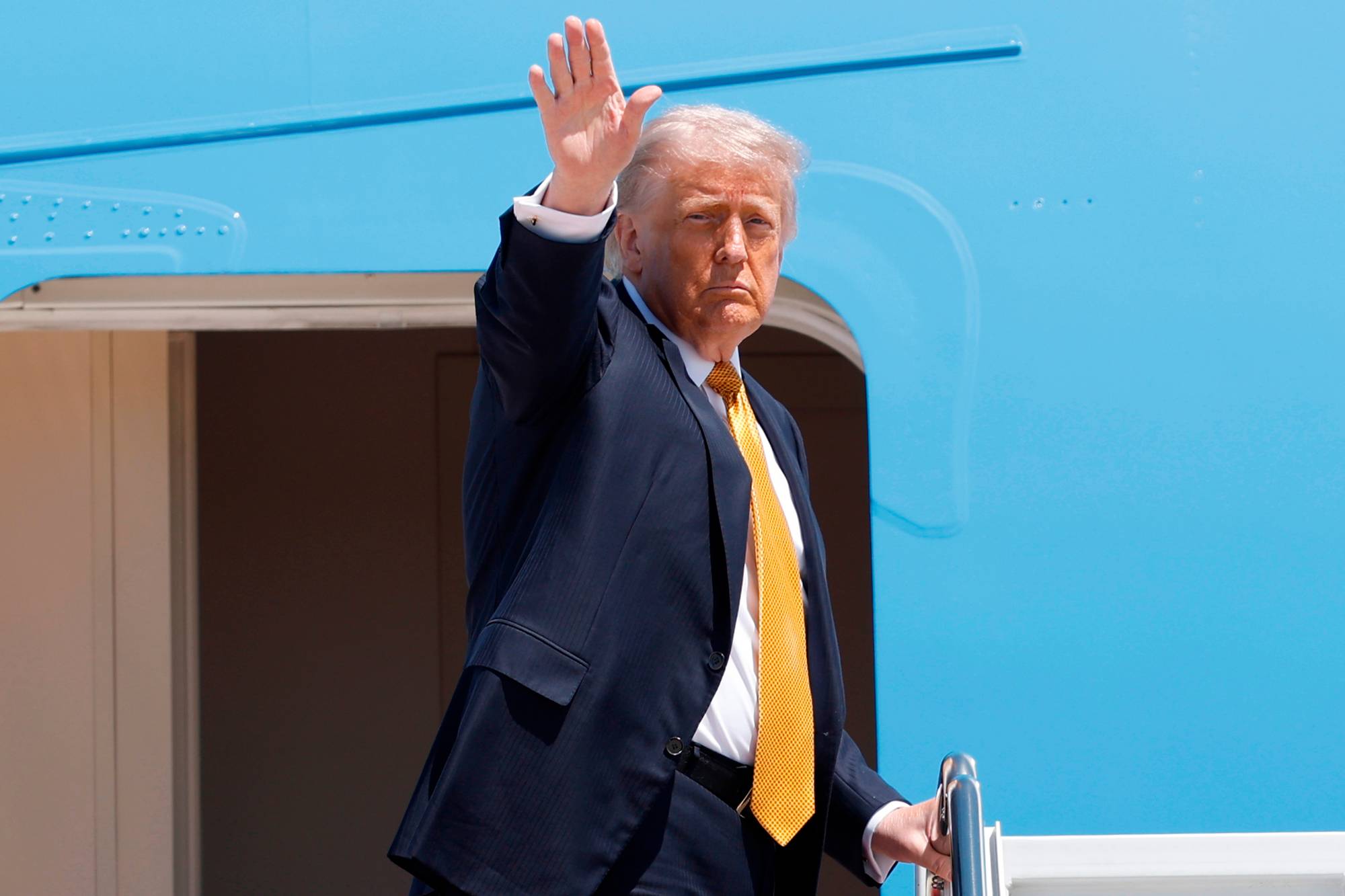 Donald Trump vinker fra et flys trapp. Han er kledd i en mørk dress og en gul slip. Trump står ved inngangen til et lyseblått fly. (Bildebeskrivelsen er laget av en KI-tjeneste)