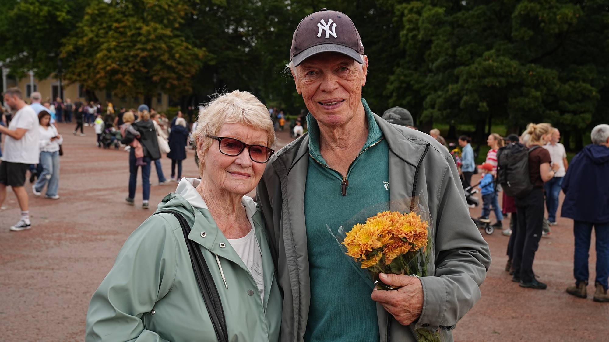 Eldre par smiler til kamera utendørs, mannen holder en blomsterbukett. Flere folk i bakgrunnen. (Bildebeskrivelsen er laget av en KI-tjeneste)