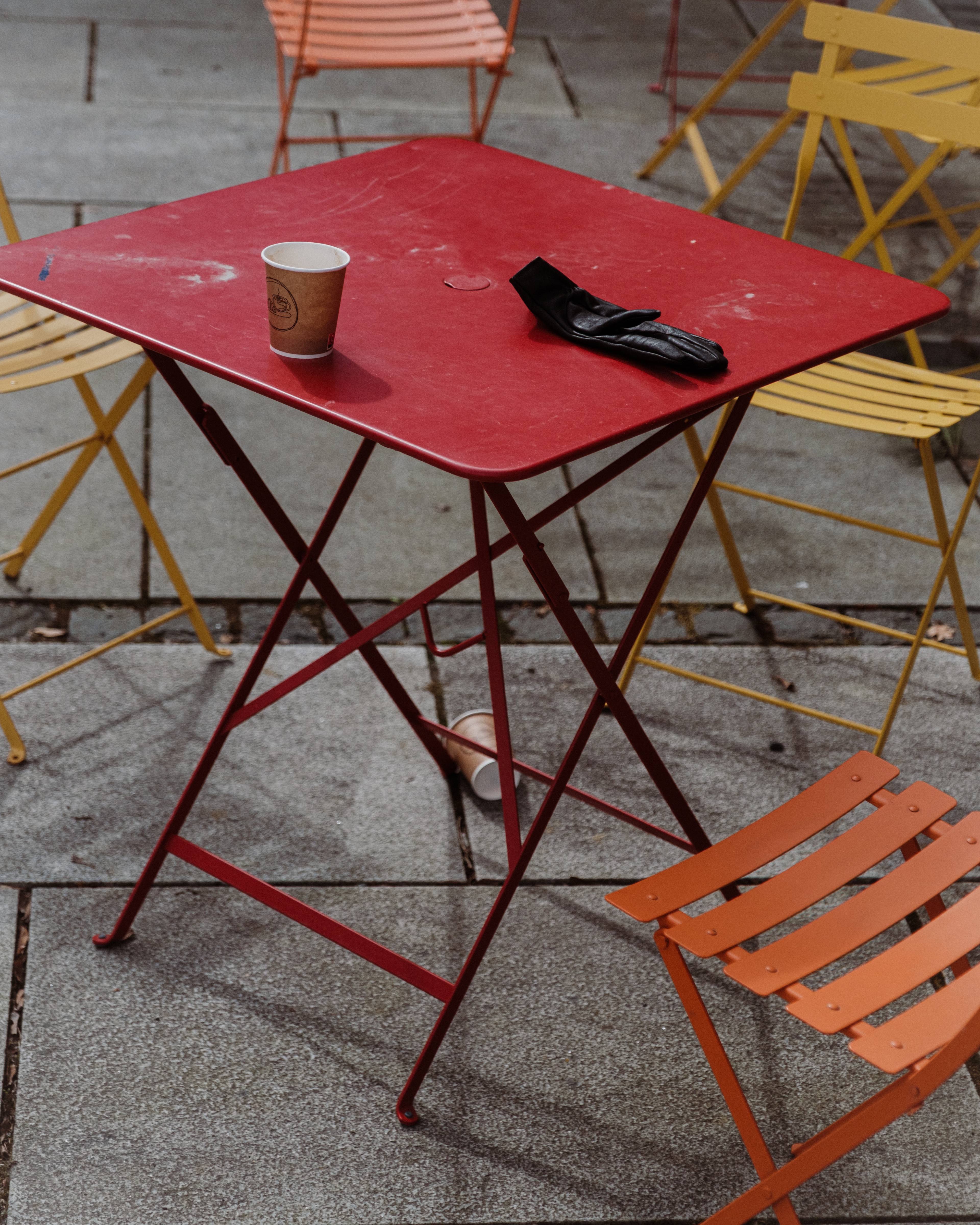 Et rødt bord, oransje og gule stoler står utendørs på en stenlagt plass. En kaffekopp og en skinnhanske ligger igjen på bordet