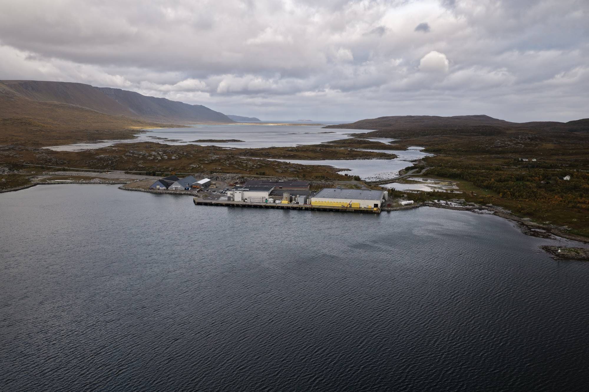Industrianlegg ved sjøen i et kupert landskap med fjell i bakgrunnen. (Bildebeskrivelsen er laget av en KI-tjeneste)