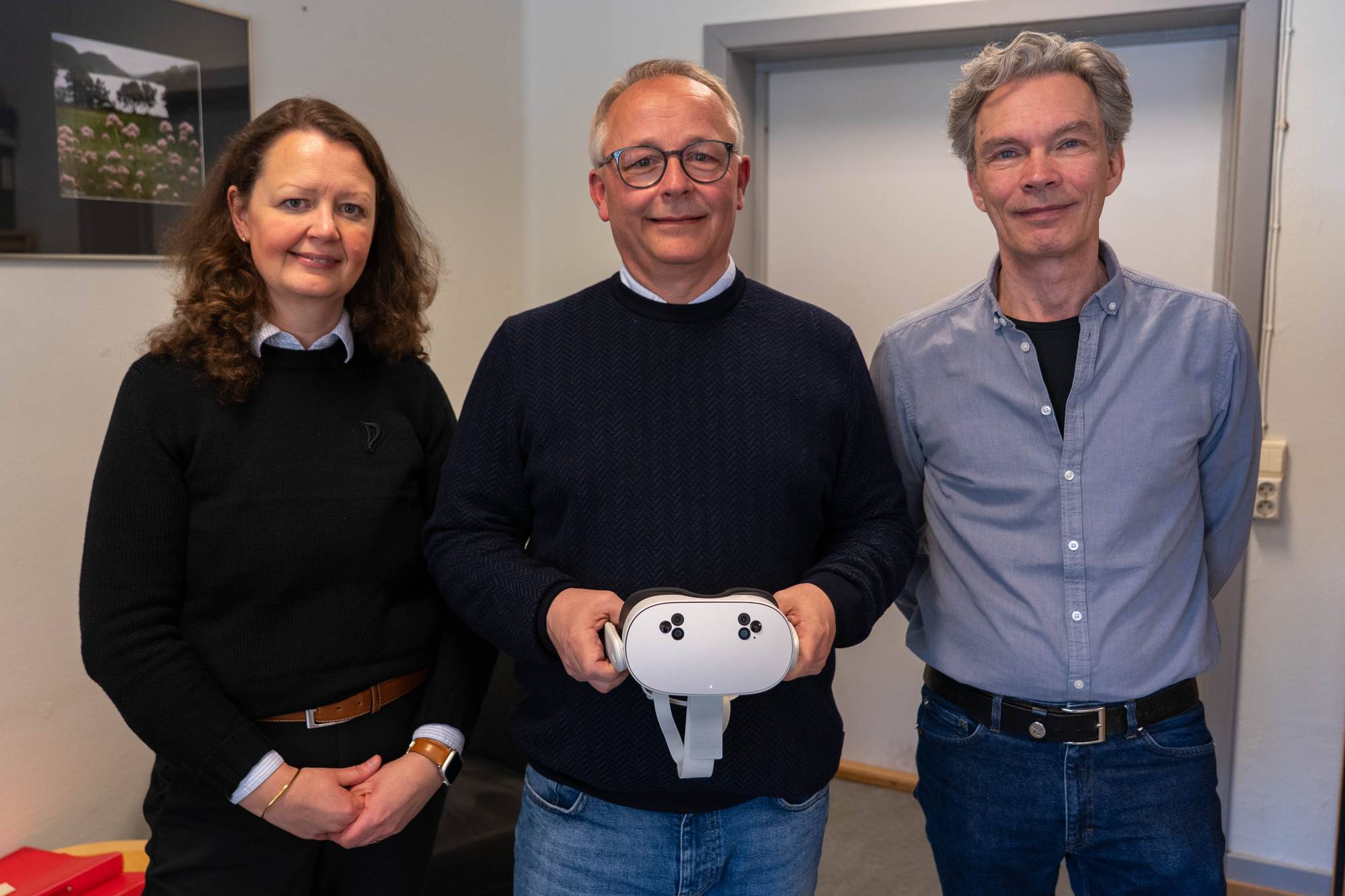 Tre personer står foran et kamera, en kvinne og to menn. Mannen i midten holder en hvit VR-brille. De står i et kontorlokale. (Bildebeskrivelsen er laget av en KI-tjeneste)