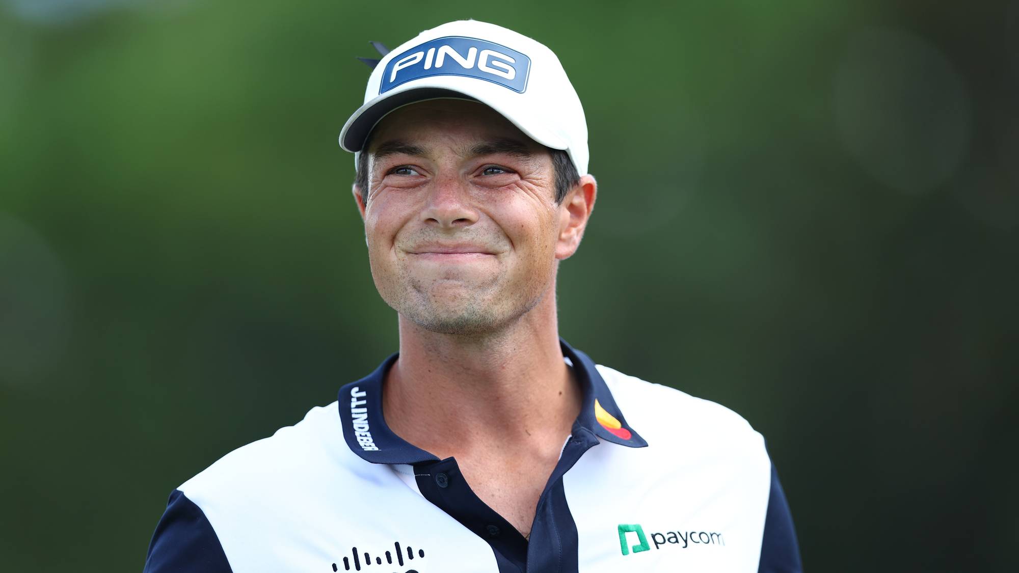 Golfspiller Viktor Hovland med caps og hvit golfskjorte smiler mot kamera. (Bildebeskrivelsen er laget av en KI-tjeneste)