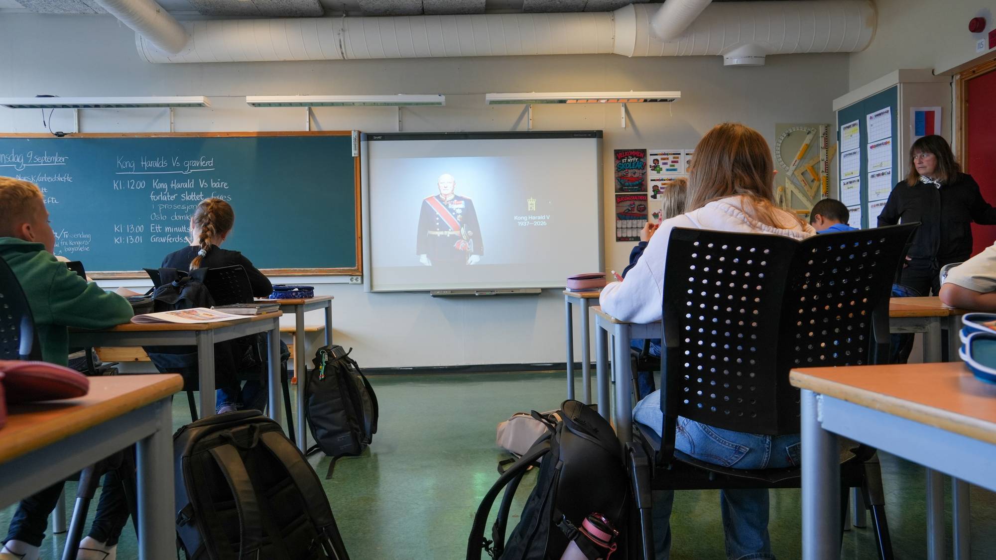 Flere elever sitter ved pultene sine i et klasserom og ser mot en skjerm som viser et bilde av kong Harald