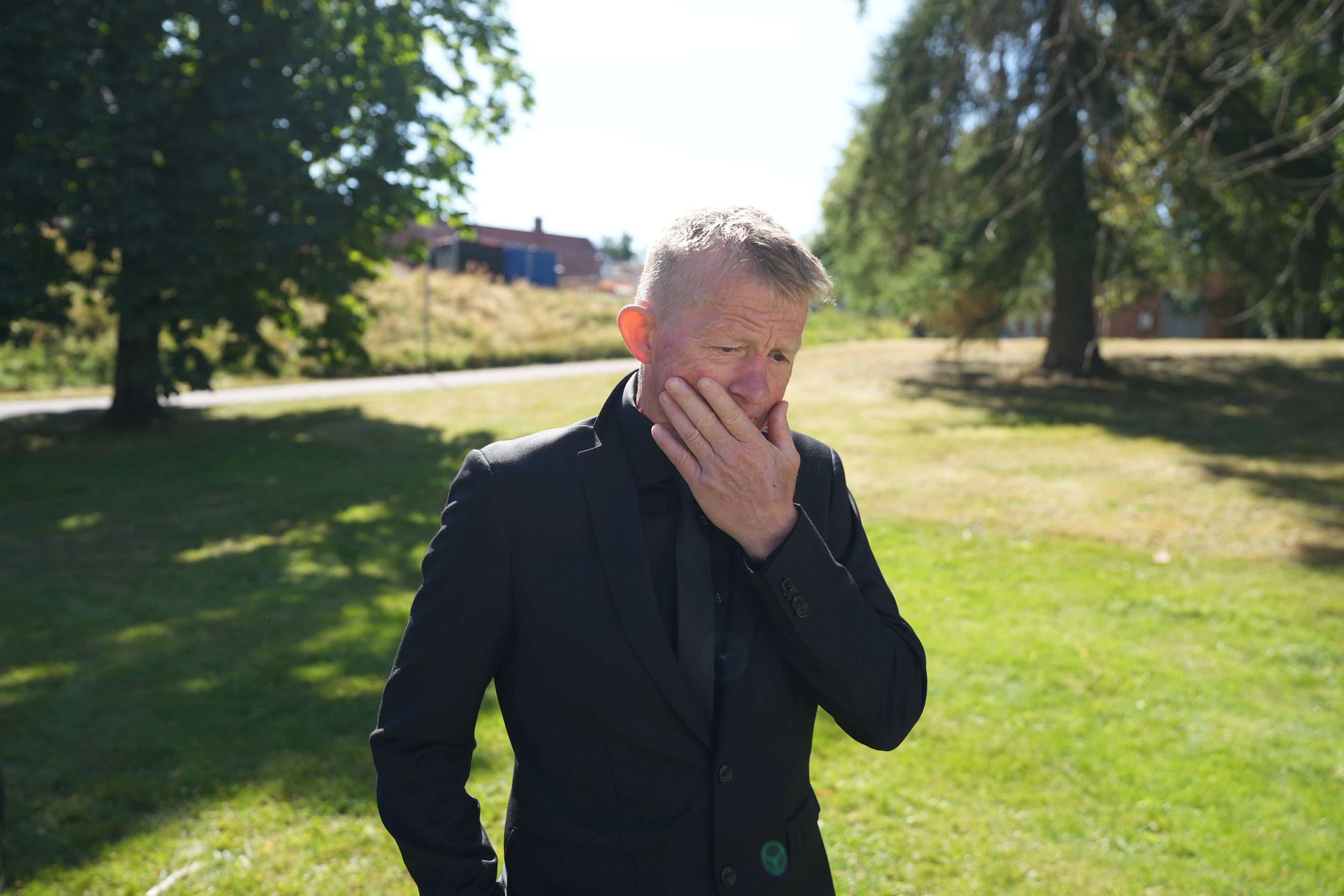 Mann i svart dress står utendørs med hånden mot ansiktet. Bakgrunnen er en park.