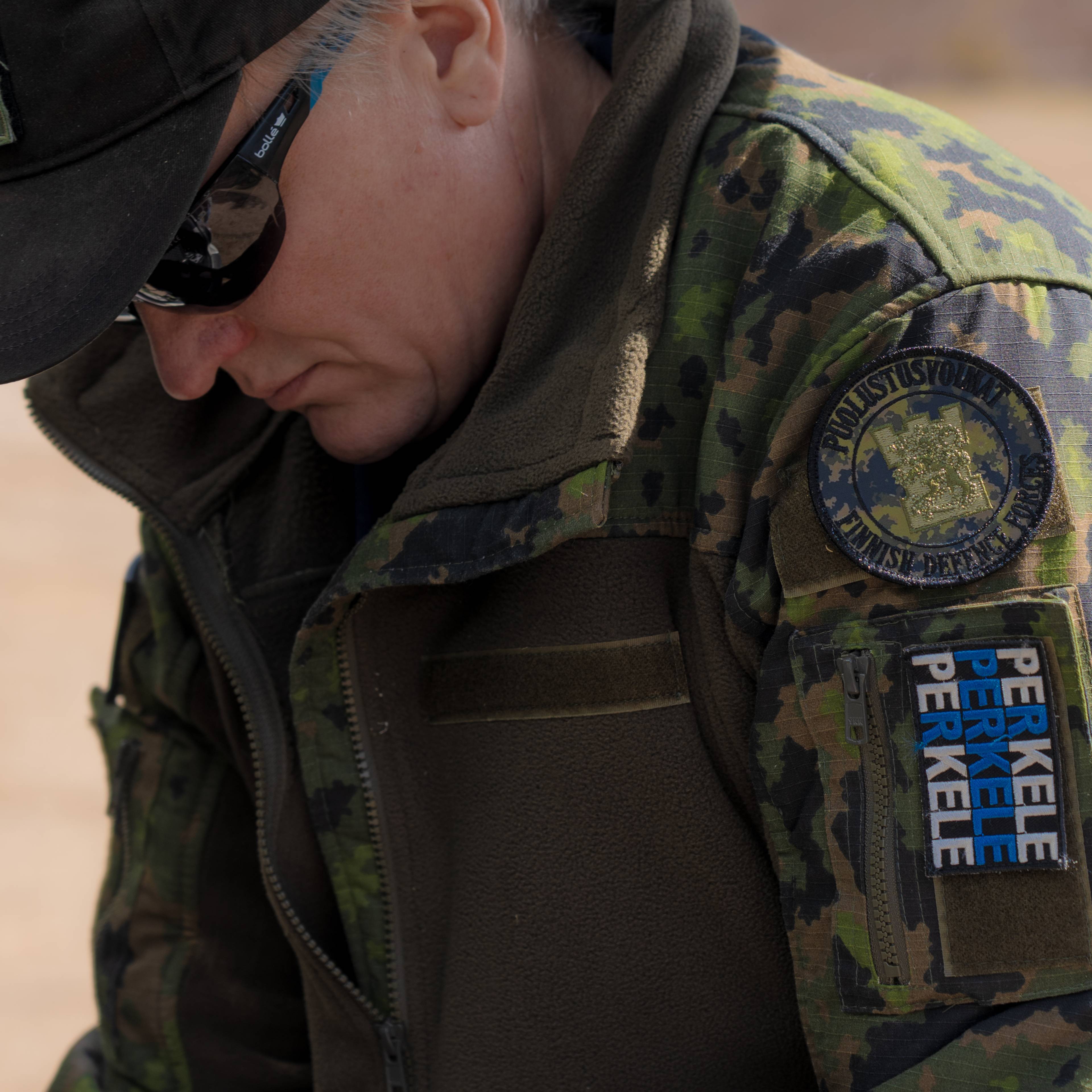 En person i kamuflasjeuniform ser ned. På uniformen er det synlige merker med tekst. Personen har på seg en caps og solbriller. Bakgrunnen er uskarp og lys. (Bildebeskrivelsen er laget av en KI-tjeneste)