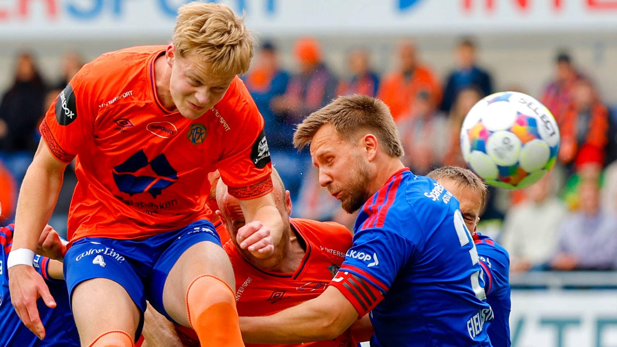 Fotballspillere i oransje og blå drakter kjemper om ballen i en duell under en kamp. (Bildebeskrivelsen er laget av en KI-tjeneste)