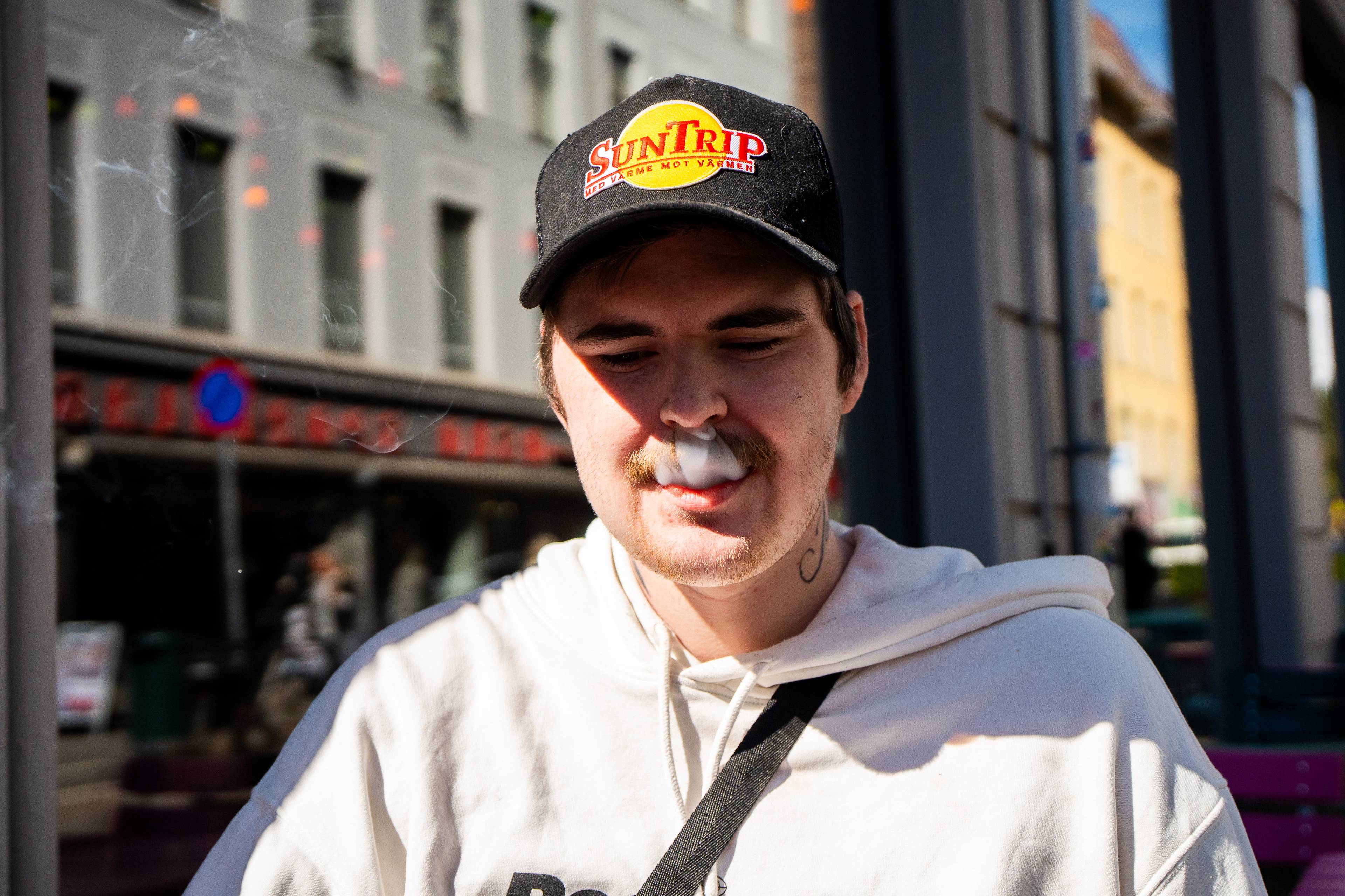 Ein mann med kaps røyker. Røyken vises som ei tåke rundt ansiktet hans. Bakgrunnen viser ei bygate med bygningar og folk. (Bildebeskrivelsen er laget av ei KI-teneste)