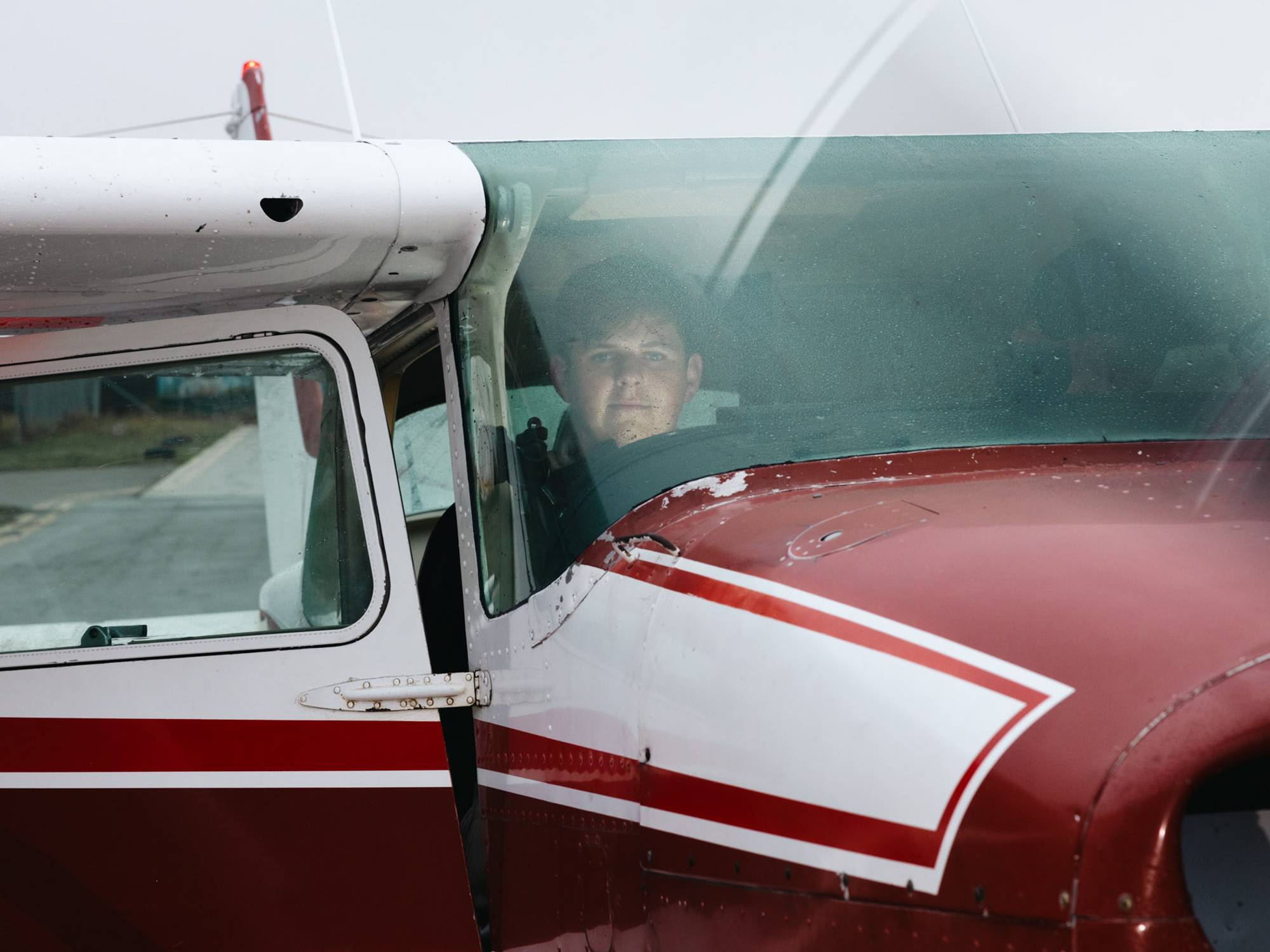 Kristoffer sitter inne i flyet, i førersetet. Han ser gjennom frontruten og inn i kamera. Han smiler. På fronten til flyet kan vi se propellen som går, han skal snart ut og fly.