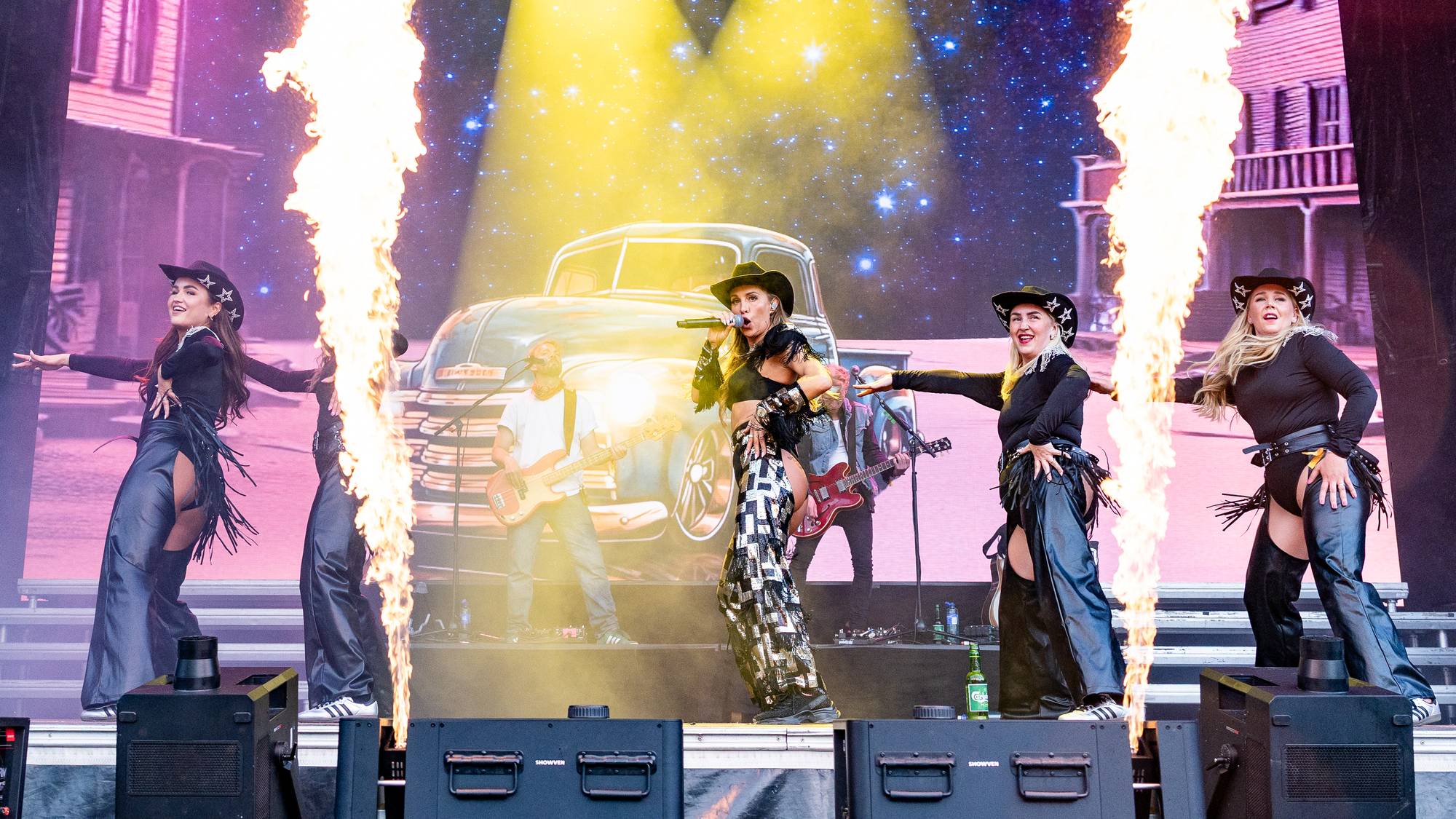 Scenen er dekorert som en westernby med flammekastere. En kvinne, kledd i cowboyhatt og matchende antrekk, synger i et headset mens hun holder en mikrofon. Rundt henne danser tre andre kvinner. En mann spiller gitar bak henne. (Bildebeskrivelsen er laget av en KI-tjeneste)